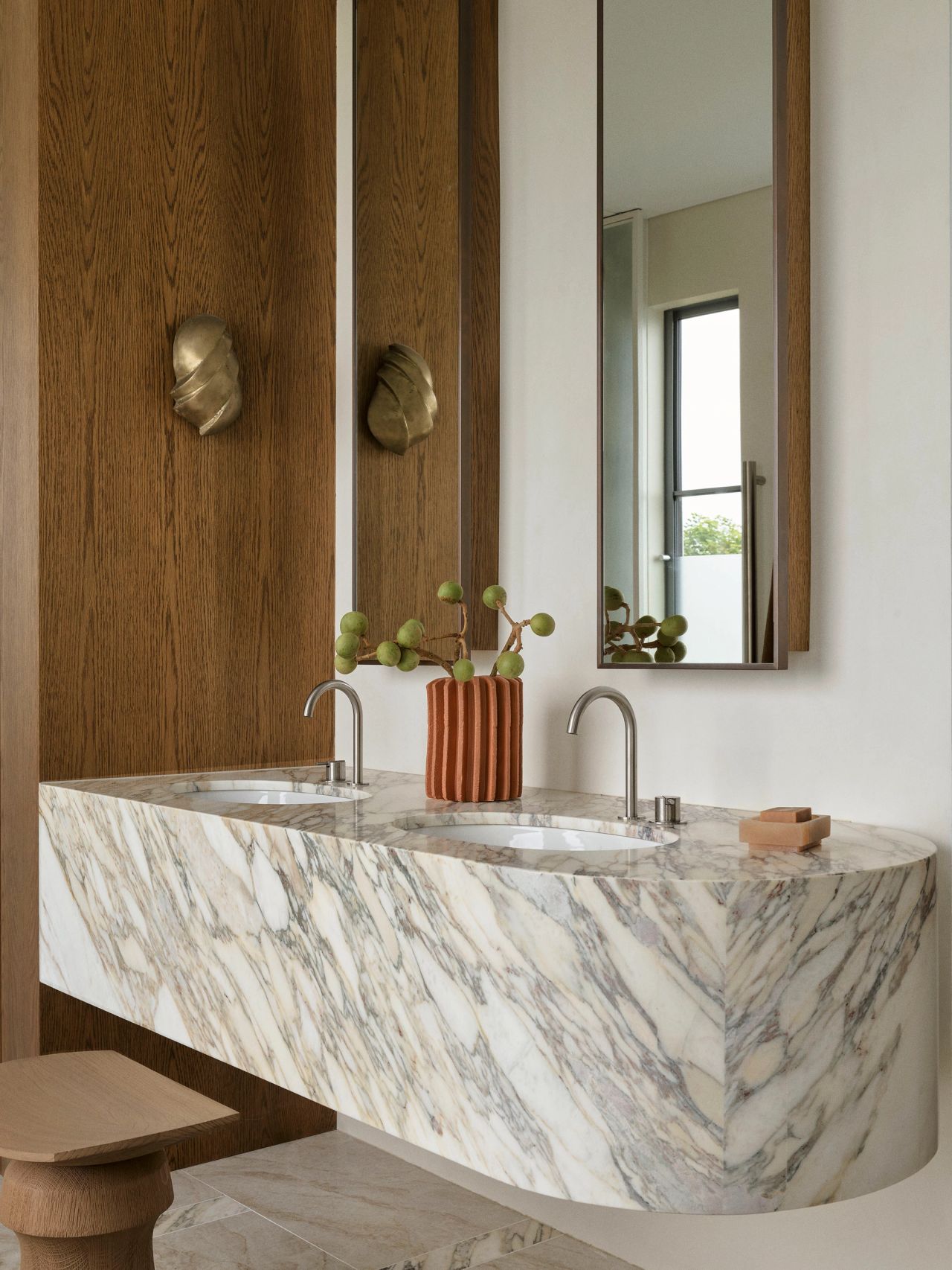 Dancing on the Ceiling Rounded double vanity in dramatic veined marble with chrome tapware, wood-framed mirrors, sculptural brass wall sconces, and a striped ceramic vase, set against oak panelling