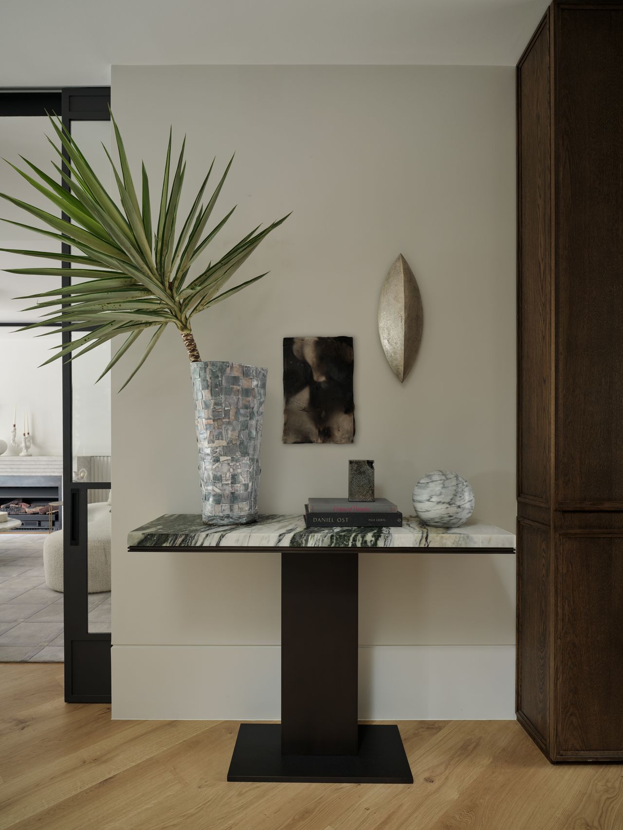 Monsieur G Entryway console table with a green-veined marble top and dark timber base, styled with a tall potted palm in a mosaic vase, a small abstract framed artwork, a leaf-shaped silver wall sculpture, a stack of books, and a marbled decorative sphere.