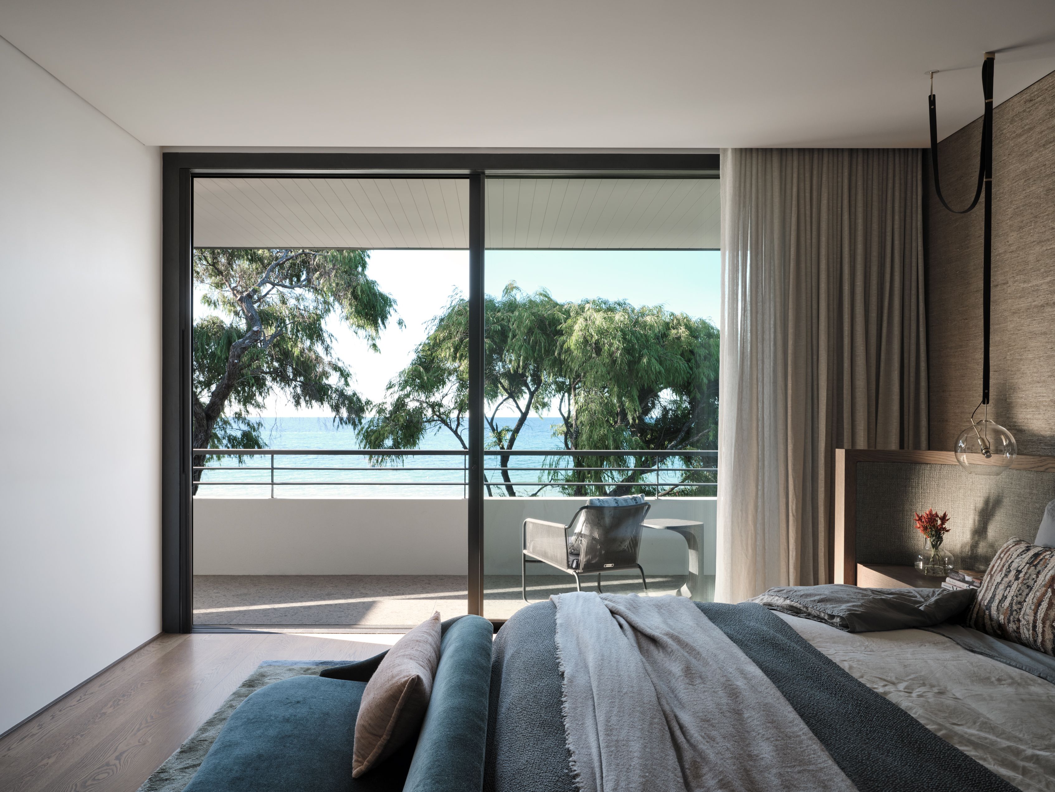 Contemporary bedroom with a timber-framed upholstered bedhead, layered grey bedding, sheer curtains and a sculptural glass pendant.