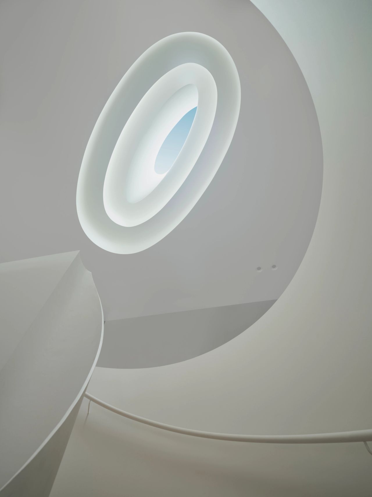 Monsieur G An abstract architectural close-up of a curved white wall featuring a deep, oval recessed skylight with concentric stepped rings, glowing softly with natural light and a glimpse of blue sky. The smooth, sculptural curve of a spiral staircase's underside sweeps through the lower portion of the frame