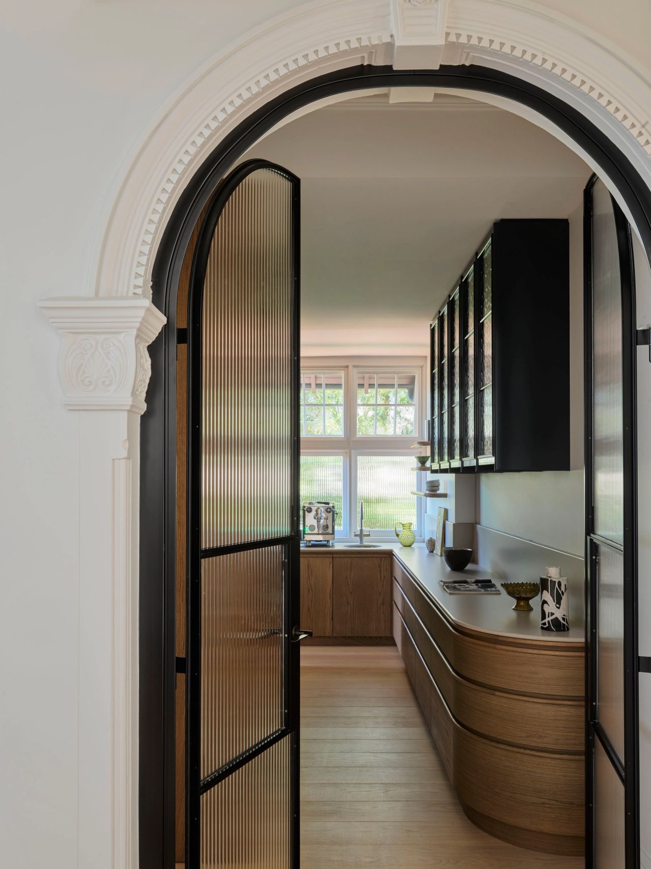 Ultramarine Ornate arched doorway with black-framed reeded glass doors opening onto a butler's pantry featuring curved wood cabinetry and dark upper cabinets with fluted glass fronts