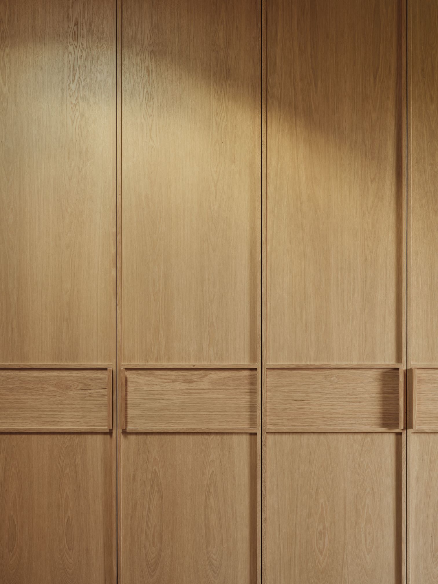 Monsieur G Detail of light oak joinery with recessed integrated handles, showing the timber grain and vertical panel divisions across a run of built-in wardrobe doors.
