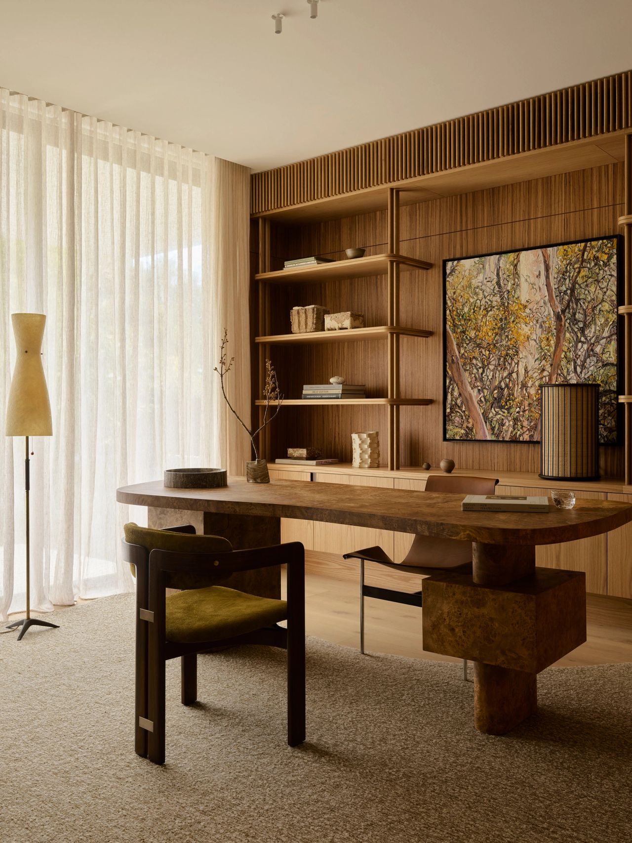 Patchwork Warm study with a burl wood desk on a sculptural pedestal base, olive velvet armchair, fluted wood shelving with styled books and objects, a tree-themed framed artwork, and sheer curtains beside an abstract floor lamp