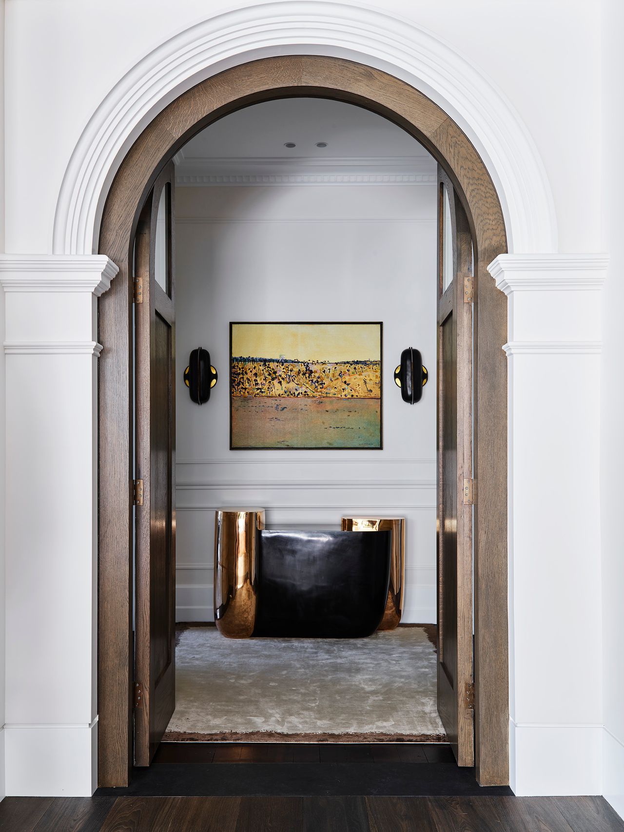 Arched timber doorway framing a refined hallway with contemporary artwork, sculptural black and brass console and wall sconces.
