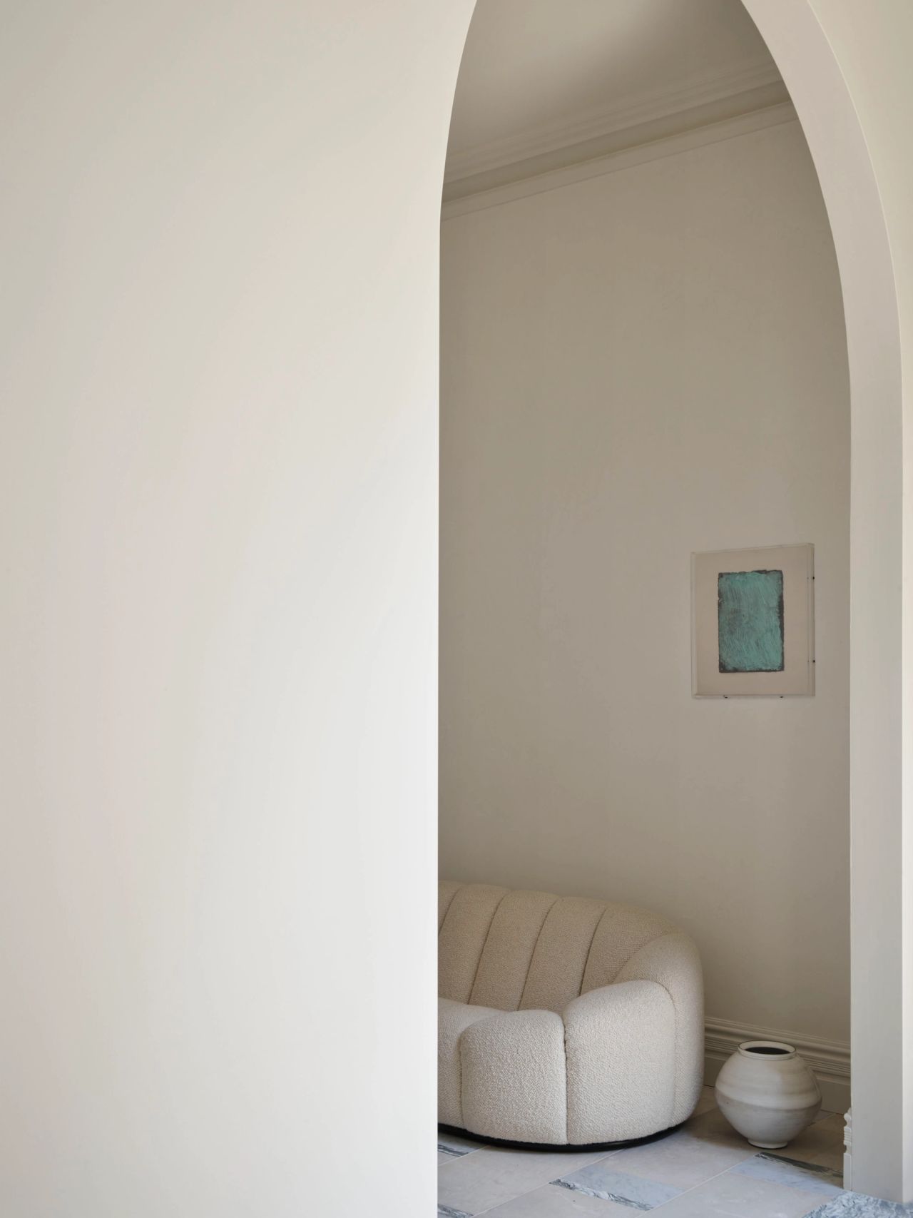 Ultramarine A minimalist alcove viewed through an arched doorway, featuring a cream boucle sofa with vertical channel-tufted cushions on a black plinth base. A small framed artwork with a muted teal-green panel hangs on the wall above, and a rounded, matte cream ceramic vase sits on the stone floor beside the sofa. Soft, even light fills the neutral, monochromatic space.