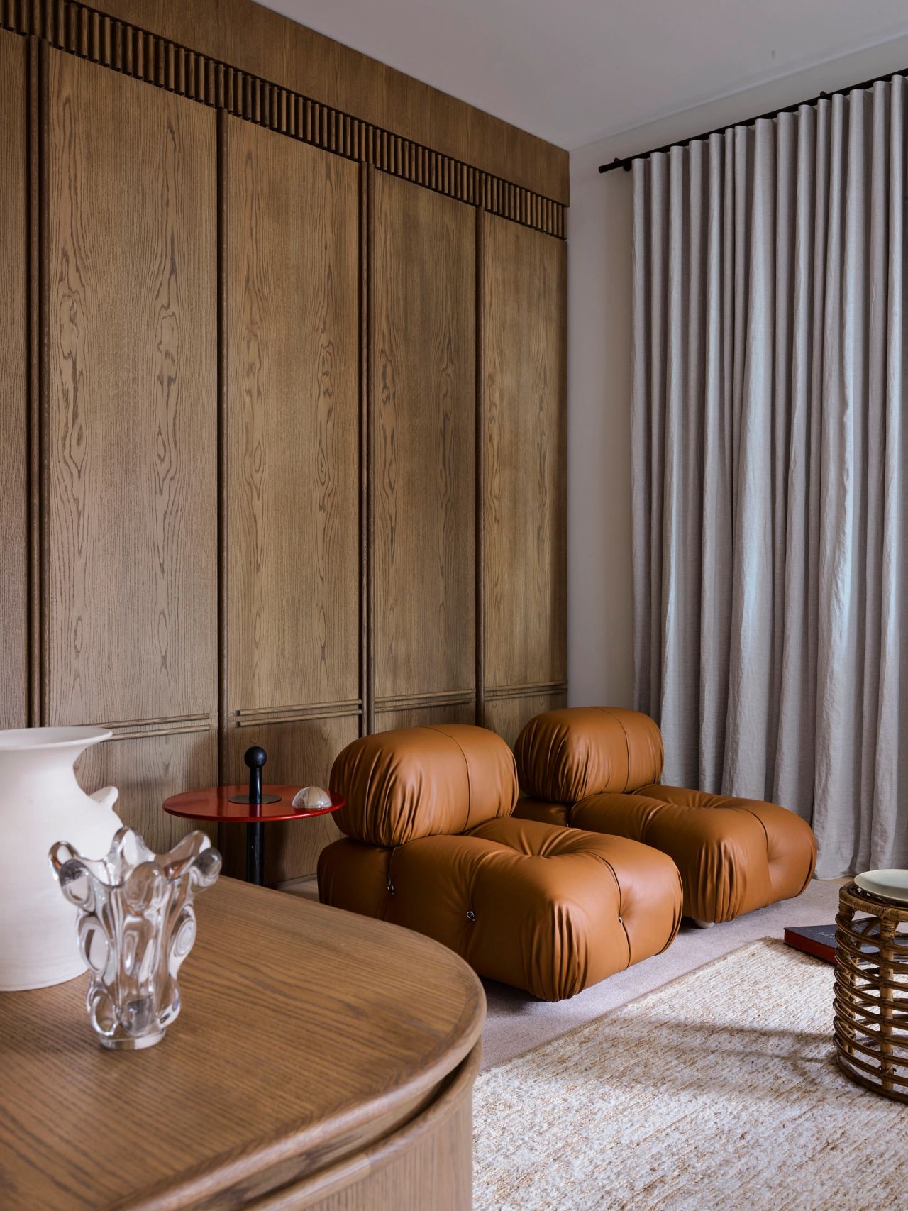 Ultramarine Camel leather modular lounge chairs against fluted wood panelling, with a small red side table, floor-to-ceiling curtains, and a glass sculptural object on a round wood table in the foreground