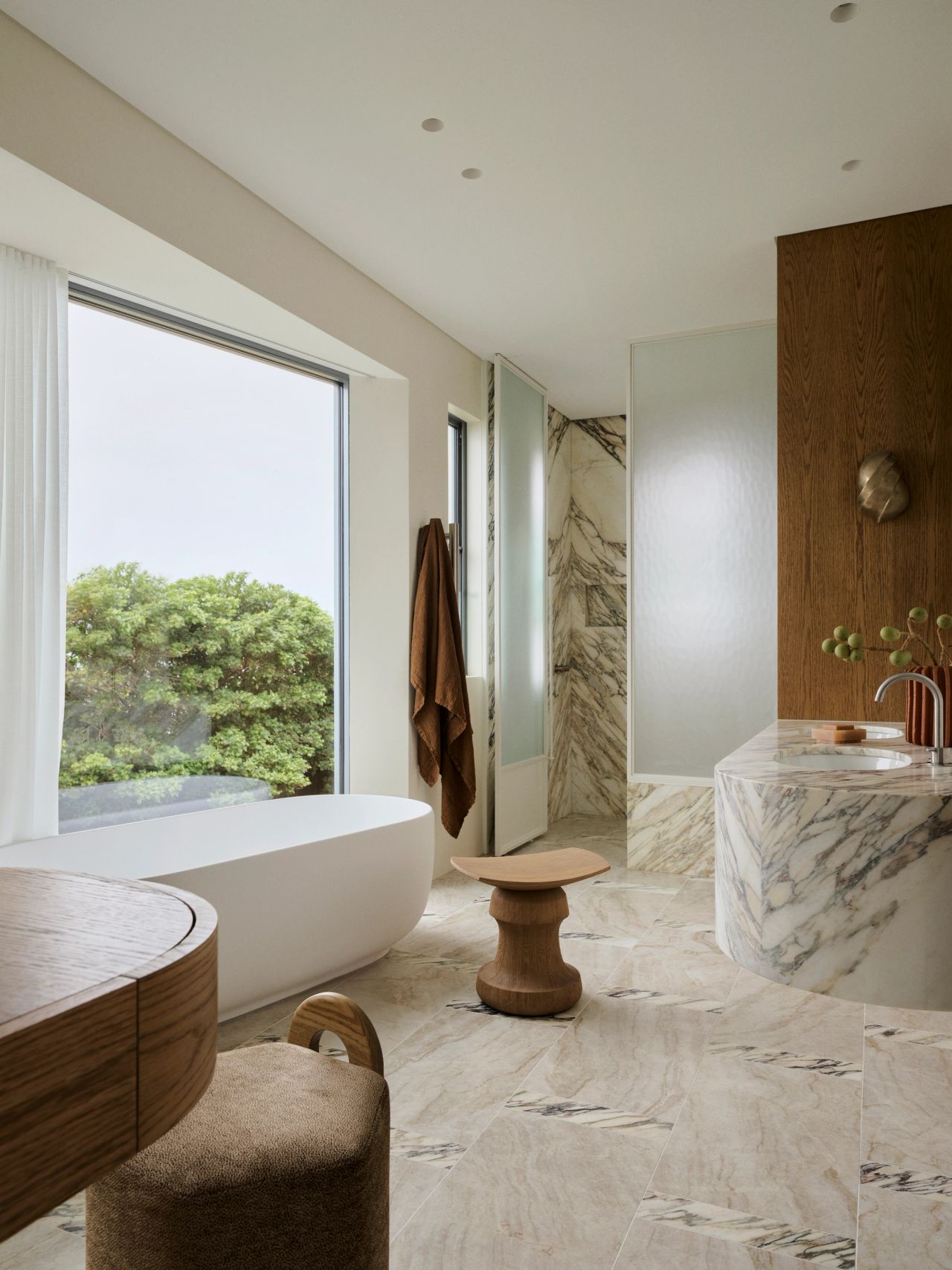 Dancing On The Ceiling Bright bathroom with a freestanding oval tub beside a large window overlooking greenery, a sculptural wooden stool, a marble vanity, and a walk-in shower with textured glass and marble walls