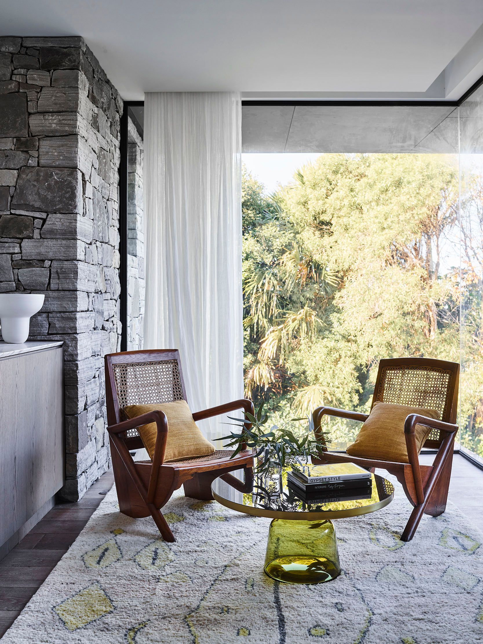 Killcare Pair of cane-back timber armchairs with mustard cushions either side of a green glass-based side table with books, set on a patterned rug beside a stacked stone wall and a garden outlook
