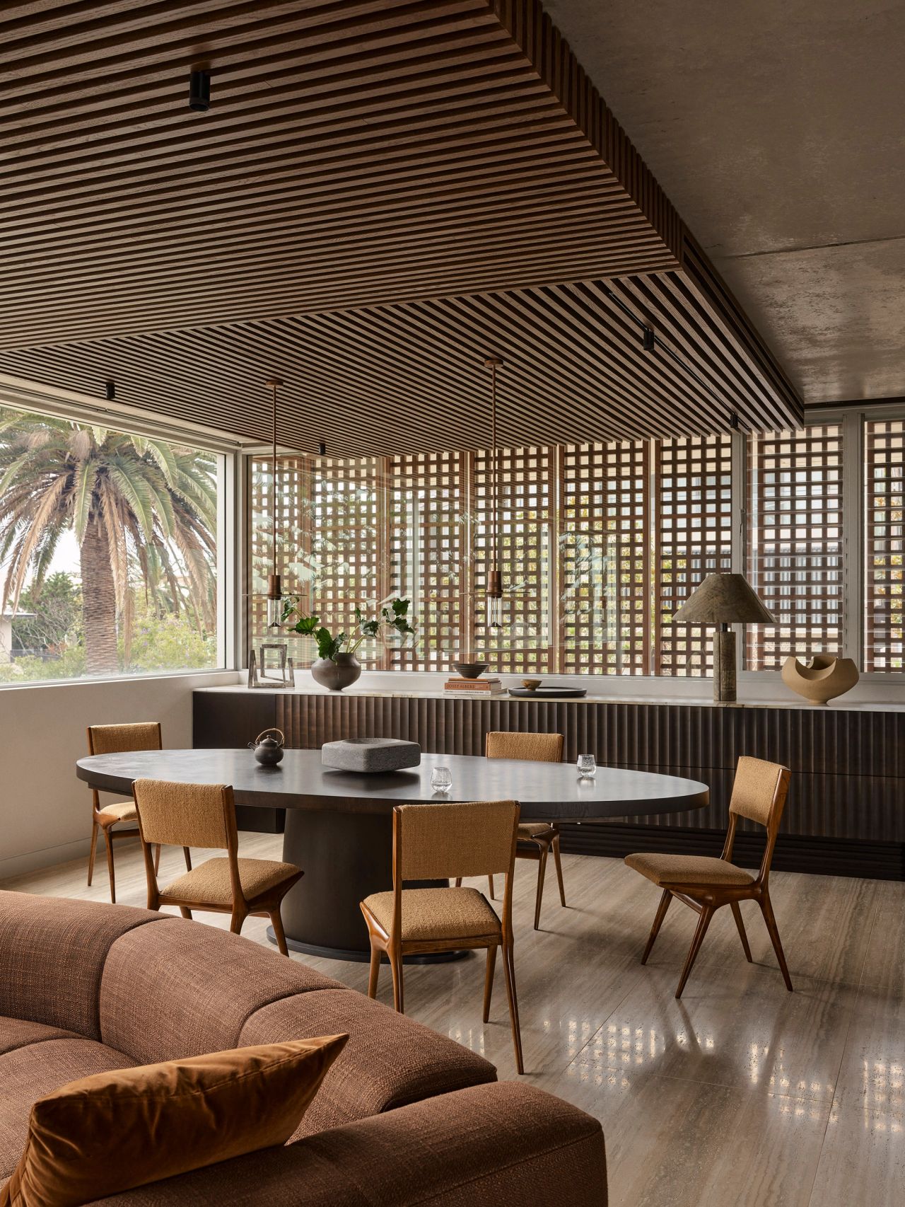 Orla Contemporary living room design open plan living and dining room blending Brazilian influences with a warm brown, black, and camel palette; black timber table with brown timber chairs. Timber batten ceiling detail and lattice timber facade treatment