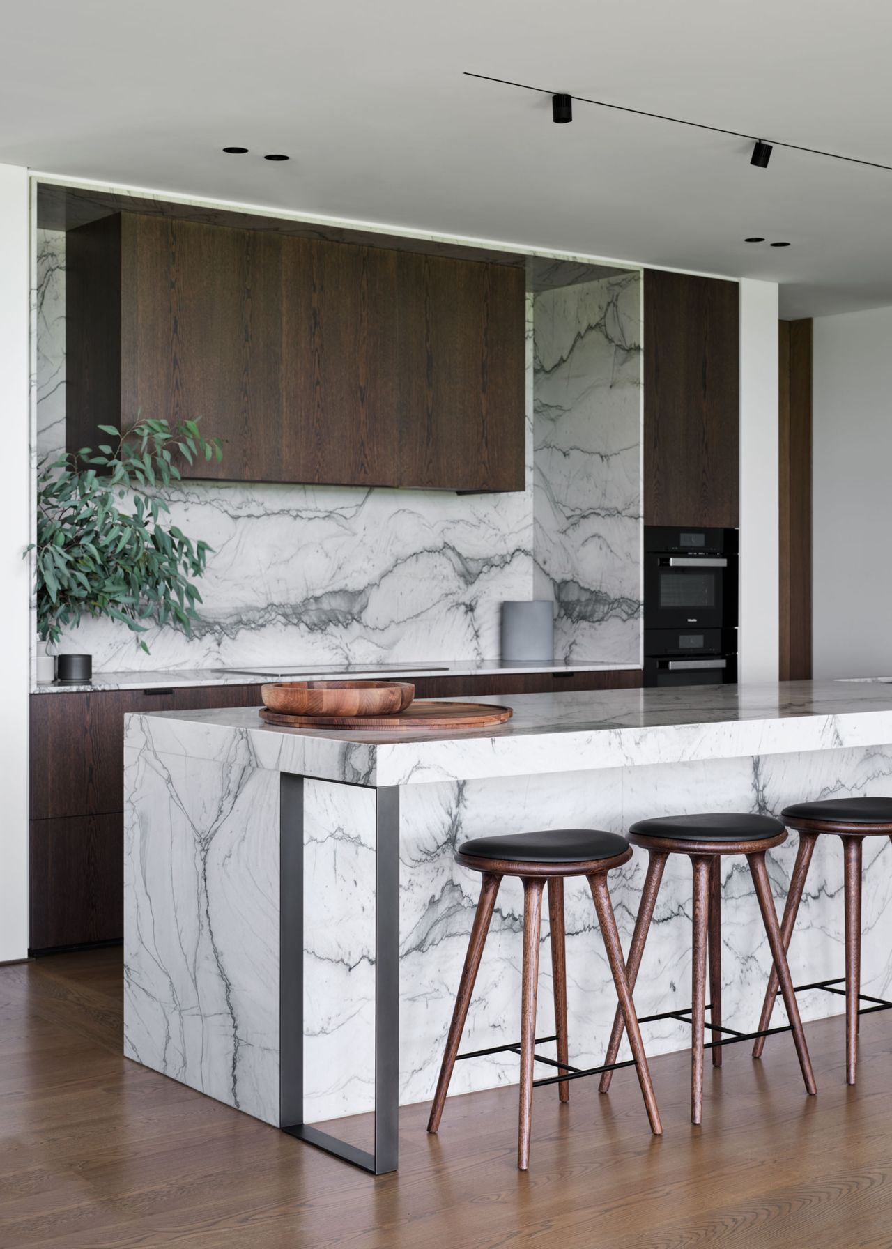 Minimalist luxury kitchen pairing richly veined white marble with dark timber cabinetry, warm wood flooring and elegant bar seating.