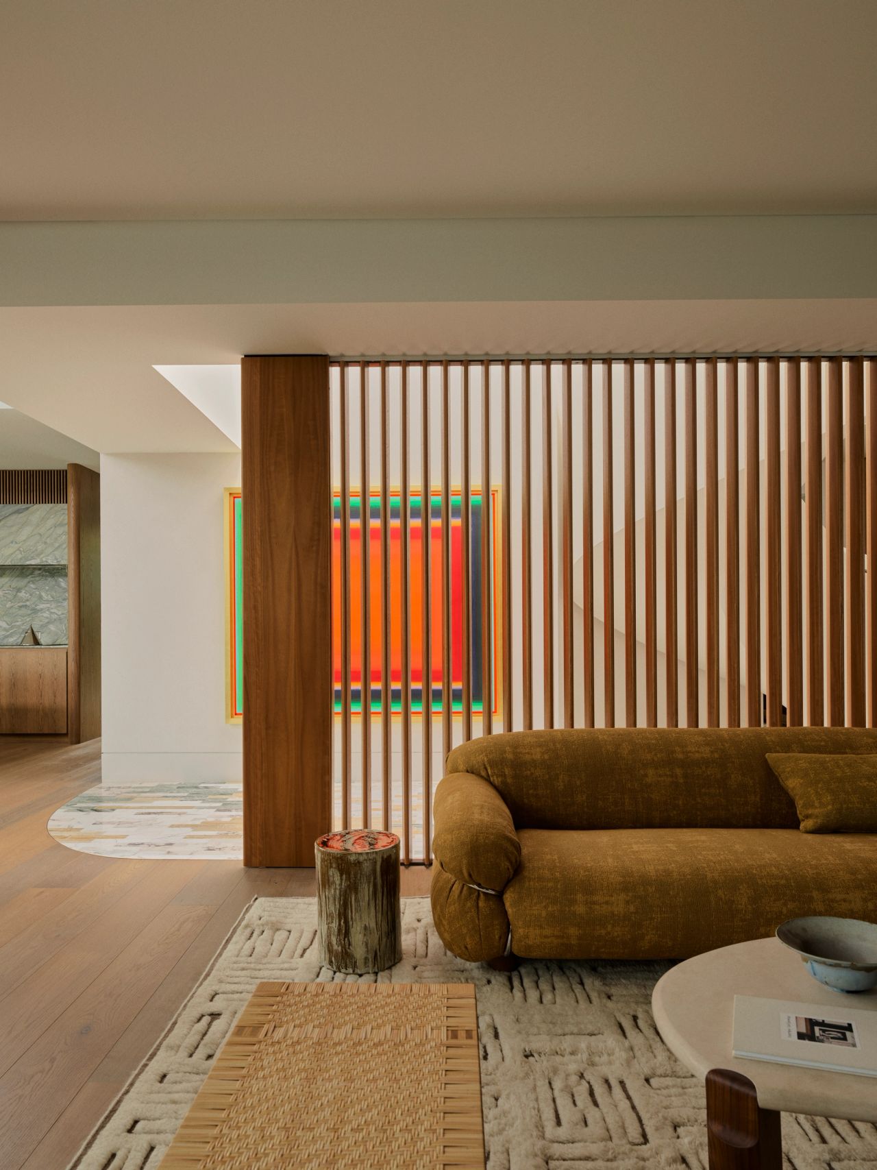 Dancing on the Ceiling Living room with an olive velvet sofa, a raw wood stump side table, and a bold geometric artwork with concentric red and orange squares, set against a vertical wood-slatted partition wall.