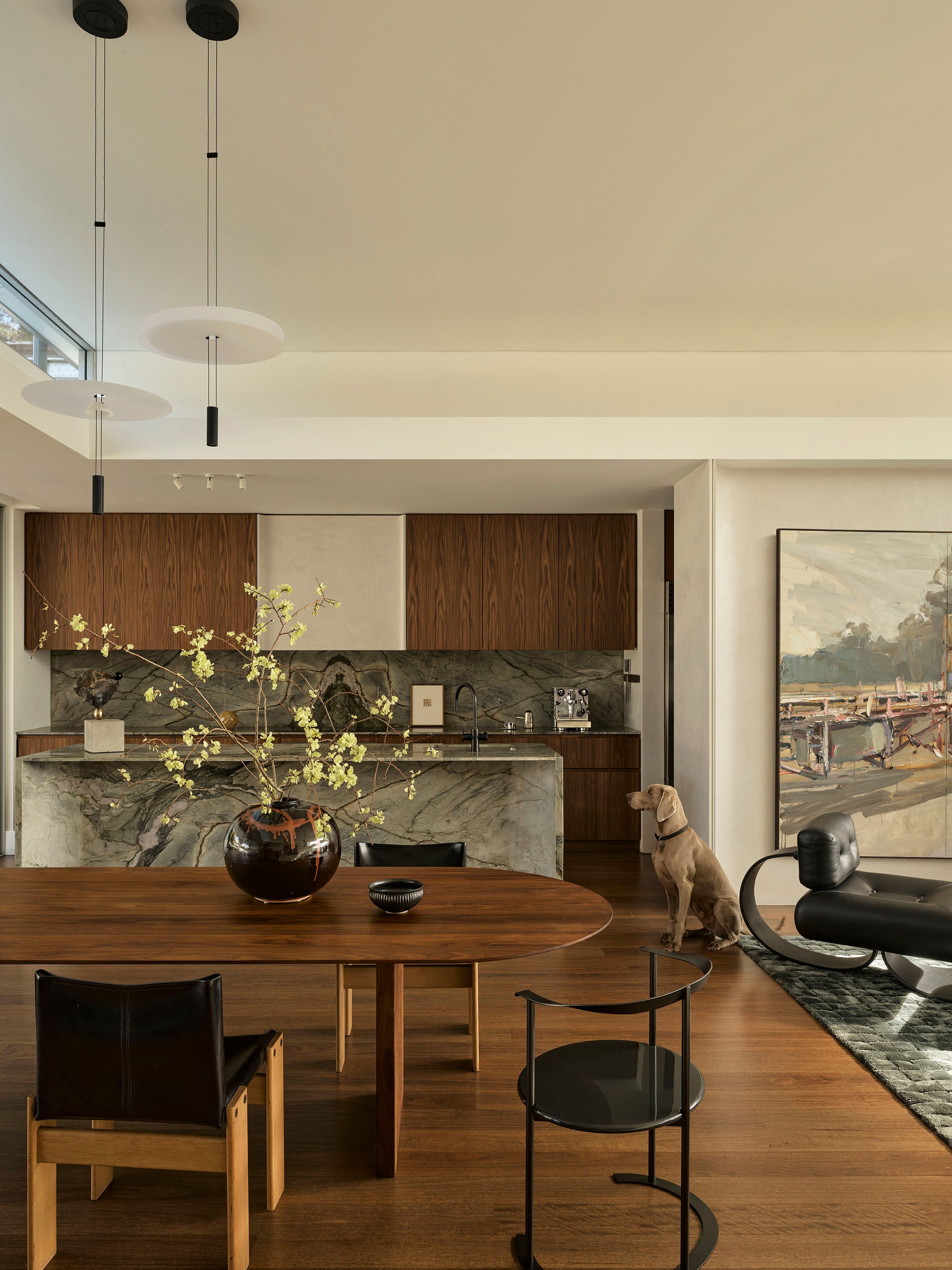 Sunset Open-plan dining and kitchen space with a round wood table, black leather dining chairs, a large ceramic vase with flowering branches, a dramatic marble kitchen island, a landscape painting, black leather lounge chairs, and a dog seated on the rug