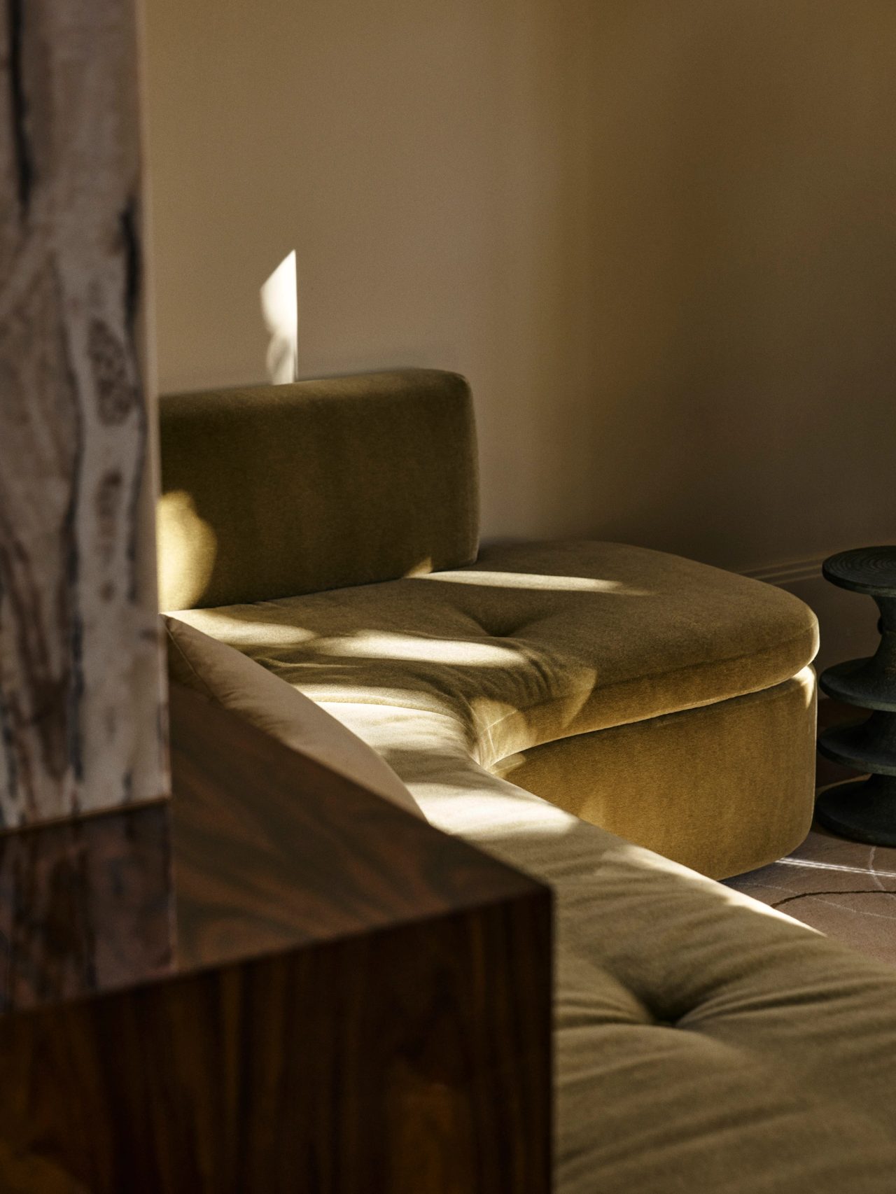 Queenie Olive velvet chaise-style sofa in raking afternoon light, with a burl wood table edge in the foreground and dark sculptural stools in the background