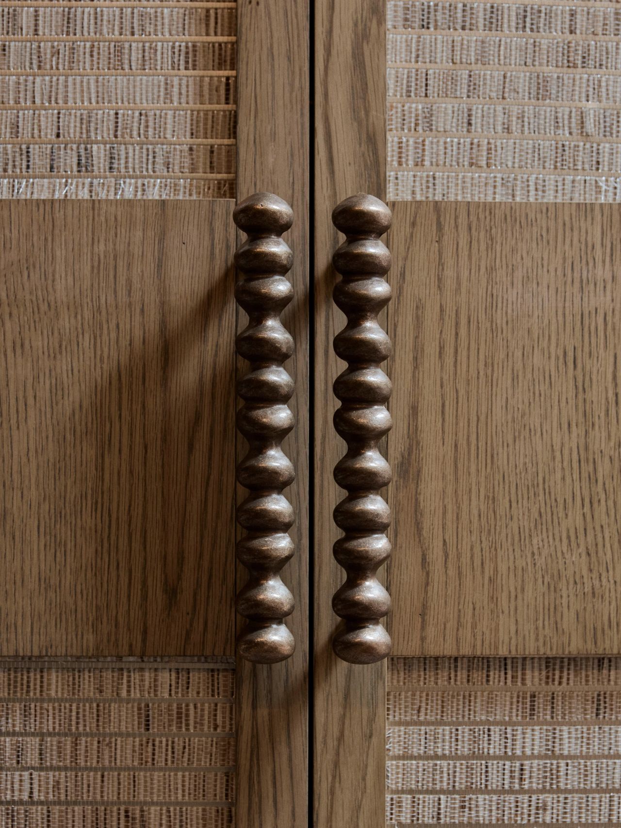 Dancing on the Ceiling Close-up of two sculptural bobbin-shaped bronze cabinet handles on wood-panelled doors bordered with woven rattan trim