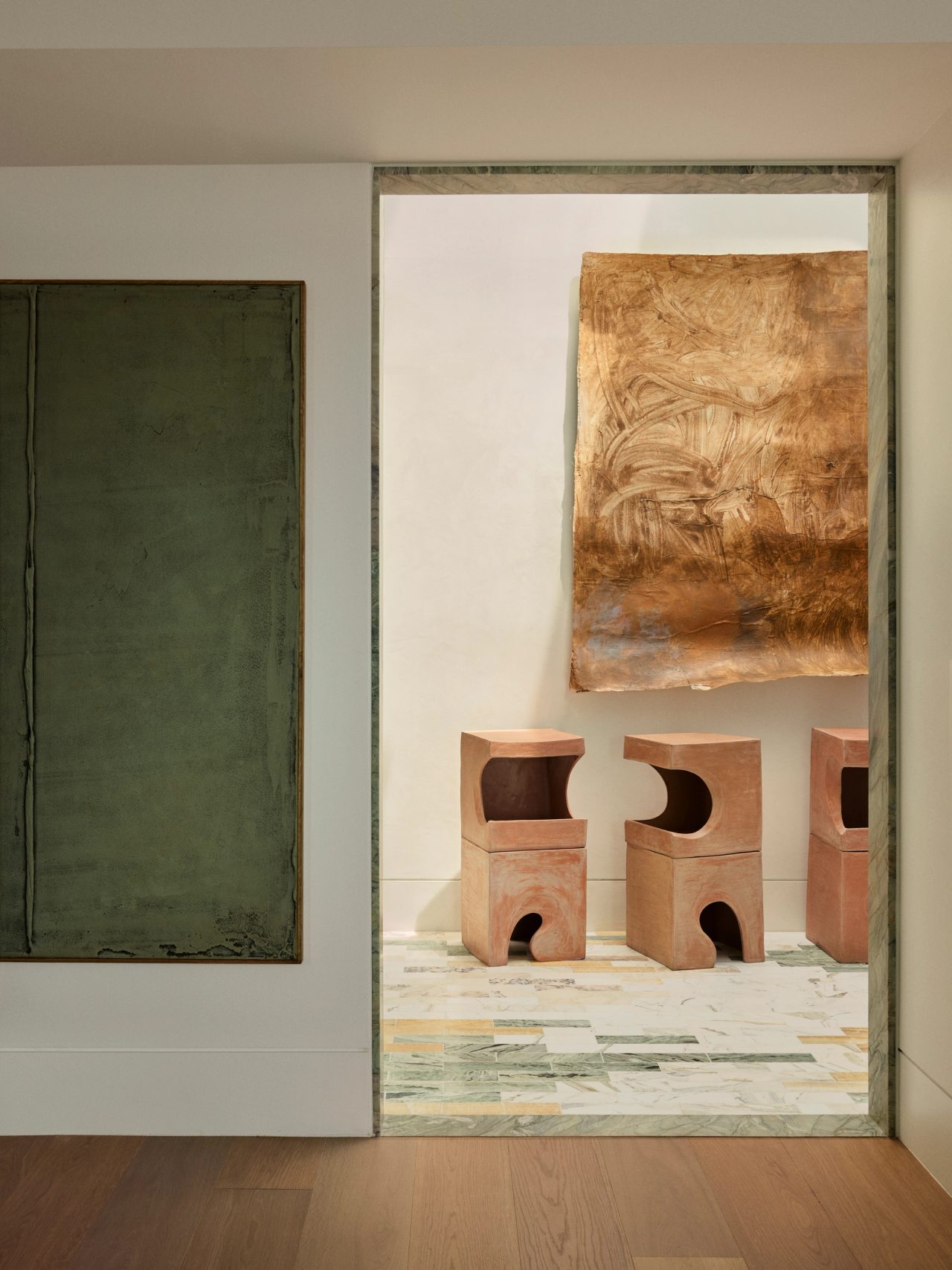 Dancing On The Ceiling Terracotta sculptural stools with geometric cutouts arranged beneath a large abstract terracotta coloured artwork, seen through a doorway framed in green stone, with a dark green artwork on the wall alongside.