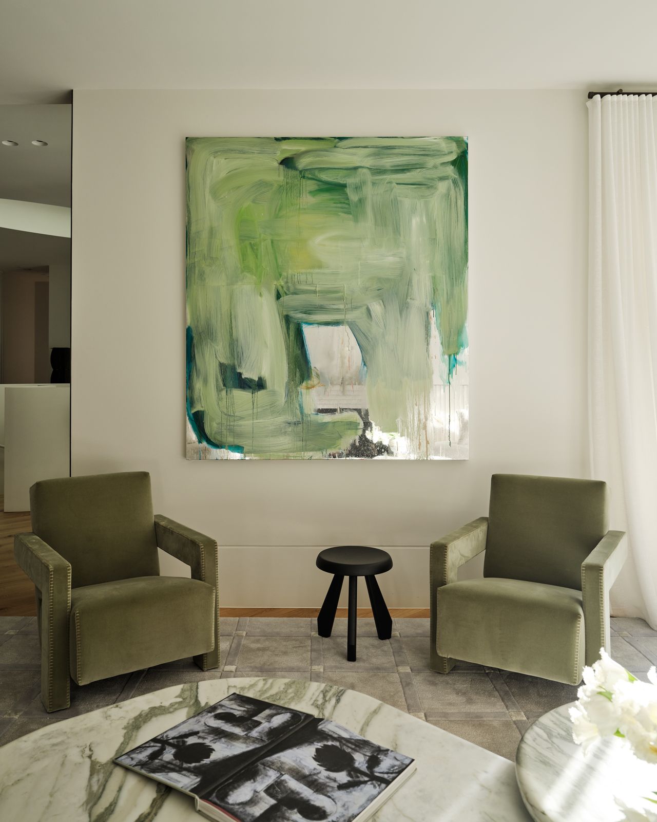 Monsieur G Sitting area with a pair of olive-green velvet armchairs flanking a black timber stool, beneath a large green abstract painting, with a marble coffee table in the foreground and sheer curtains to the side