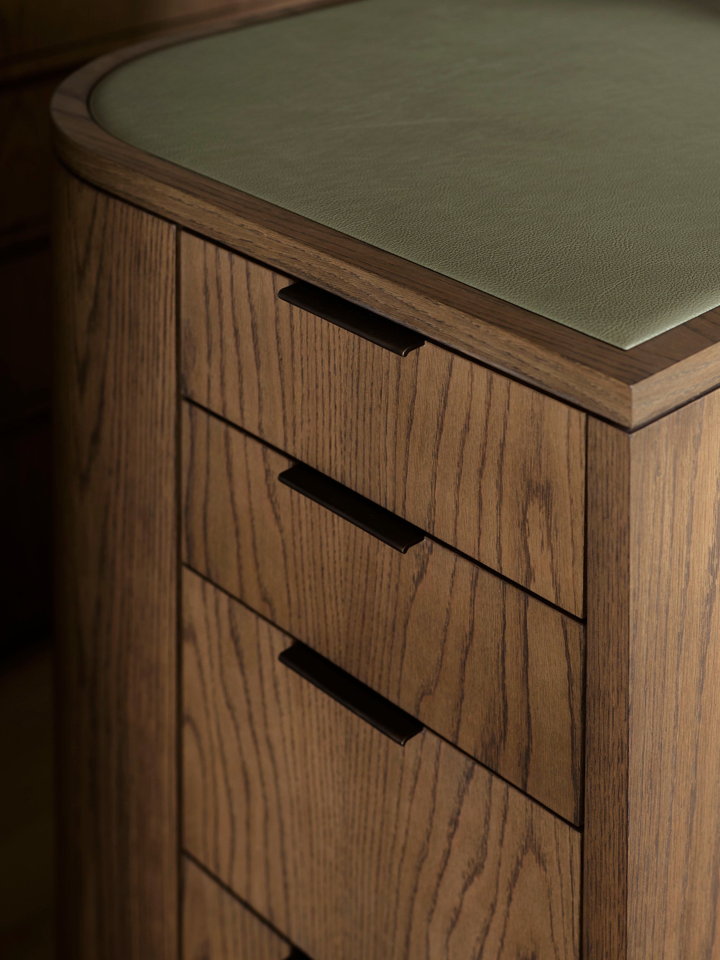 Patchwork Close-up of a rounded wooden desk corner with a sage green leather inset top and slim brass drawer pulls on the oak drawer fronts.
