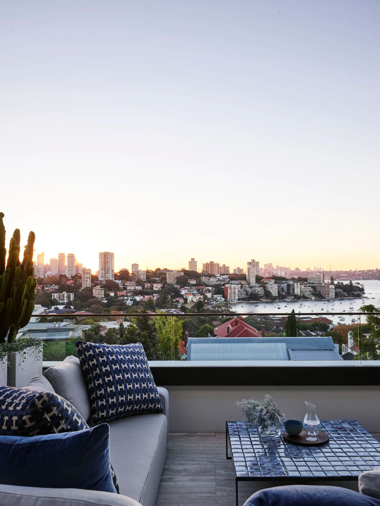 Contemporary balcony lounge with blue patterned cushions overlooking panoramic harbour and city skyline views at sunset.