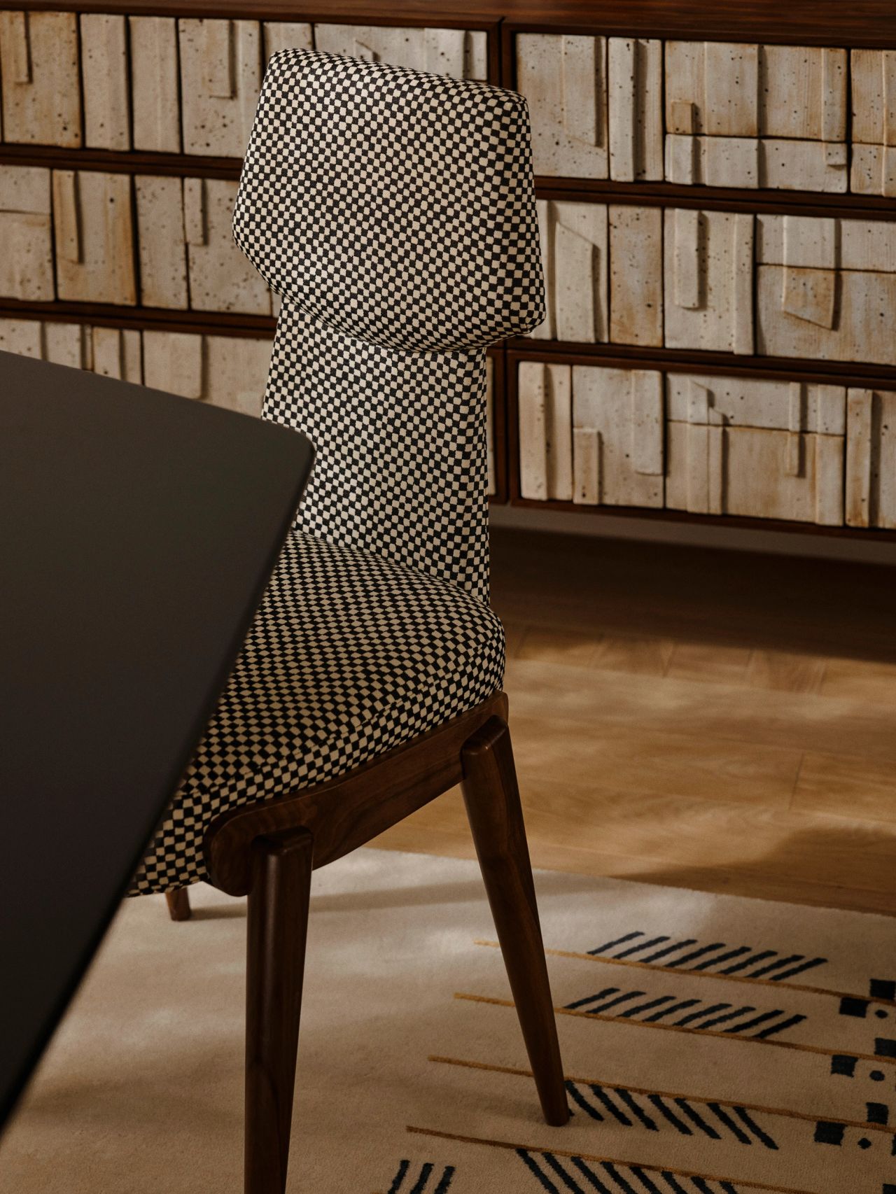 Queenie Upholstered dining chair in a black-and-cream houndstooth-style fabric with dark wood legs, set against a patterned striped rug and a textured woven-panel backdrop.