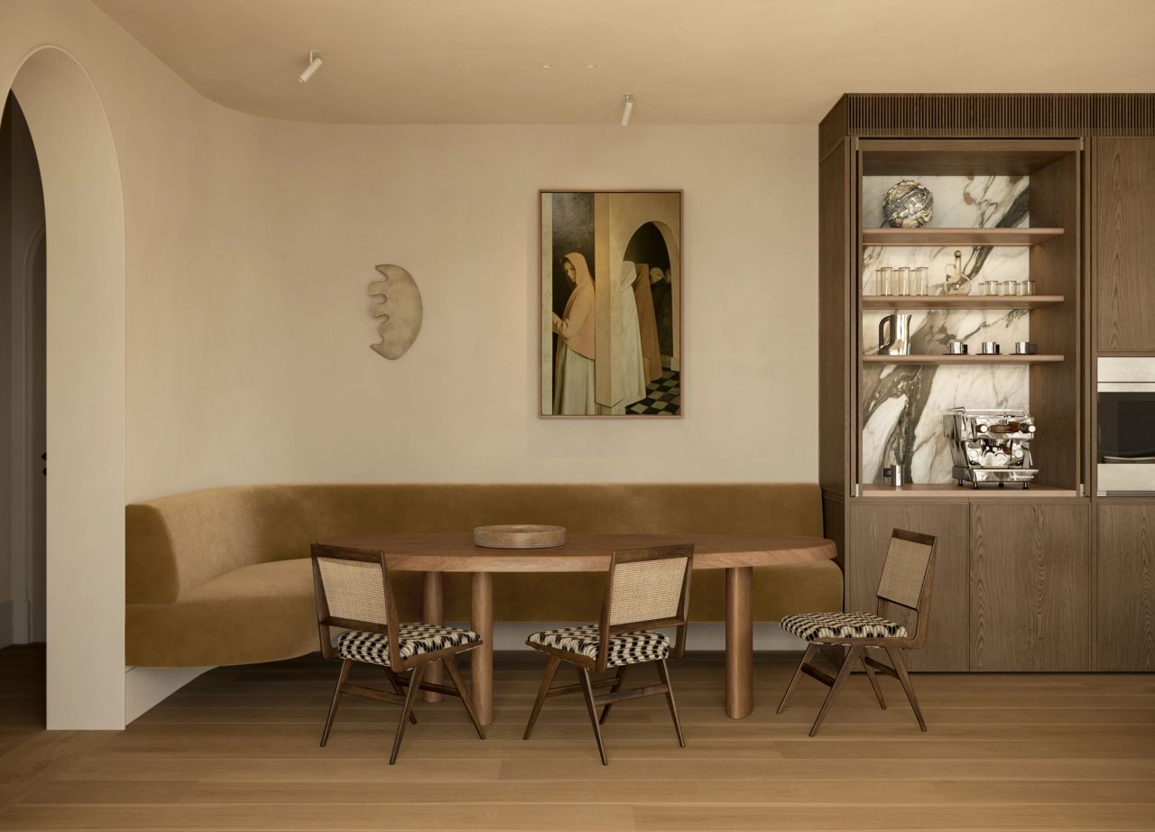 Lady M Dining nook with a built-in ochre banquette, oval timber table, and cane-back chairs upholstered in a graphic black-and-white weave, set against a marble-lined display cabinet and a figurative painting on the wall