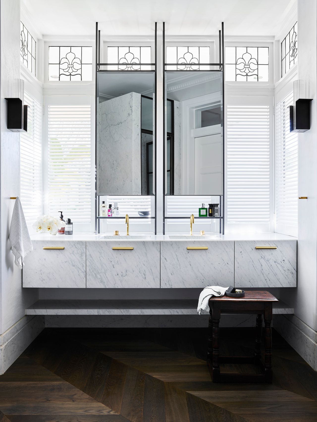 Luxurious double vanity with white marble cabinetry, brass fixtures and suspended mirrors framed by decorative windows.