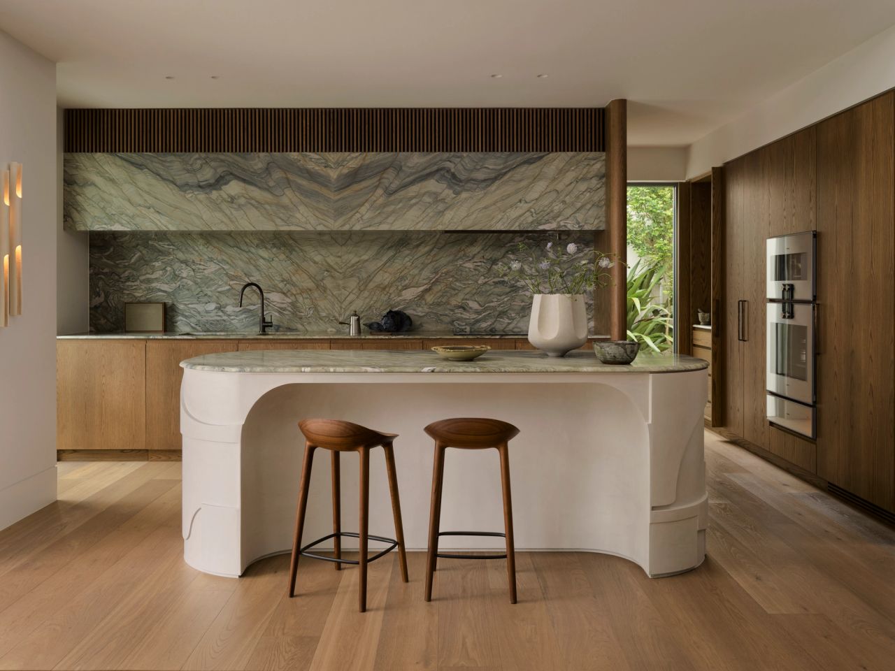 Dancing On The Ceiling Sculptural white plaster kitchen island with a book-matched marble top, paired with two dark wood bar stools, set against a dramatic mirrored marble splashback and oak cabinetry