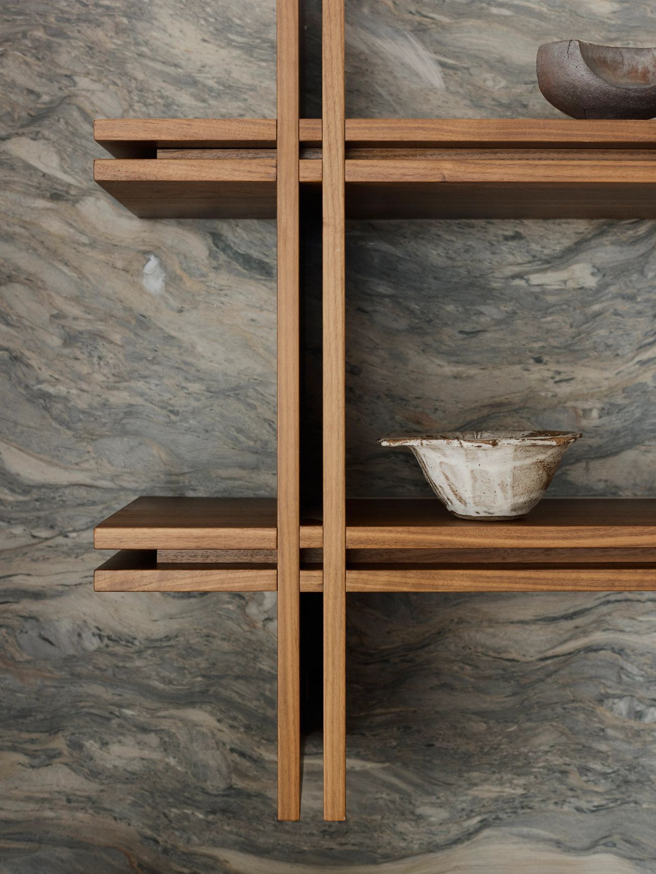 Alt text: "Orla wall-mounted walnut timber shelf with a crossed intersection detail, set against a blue-and-beige stone wall and styled with a ceramic bowl.