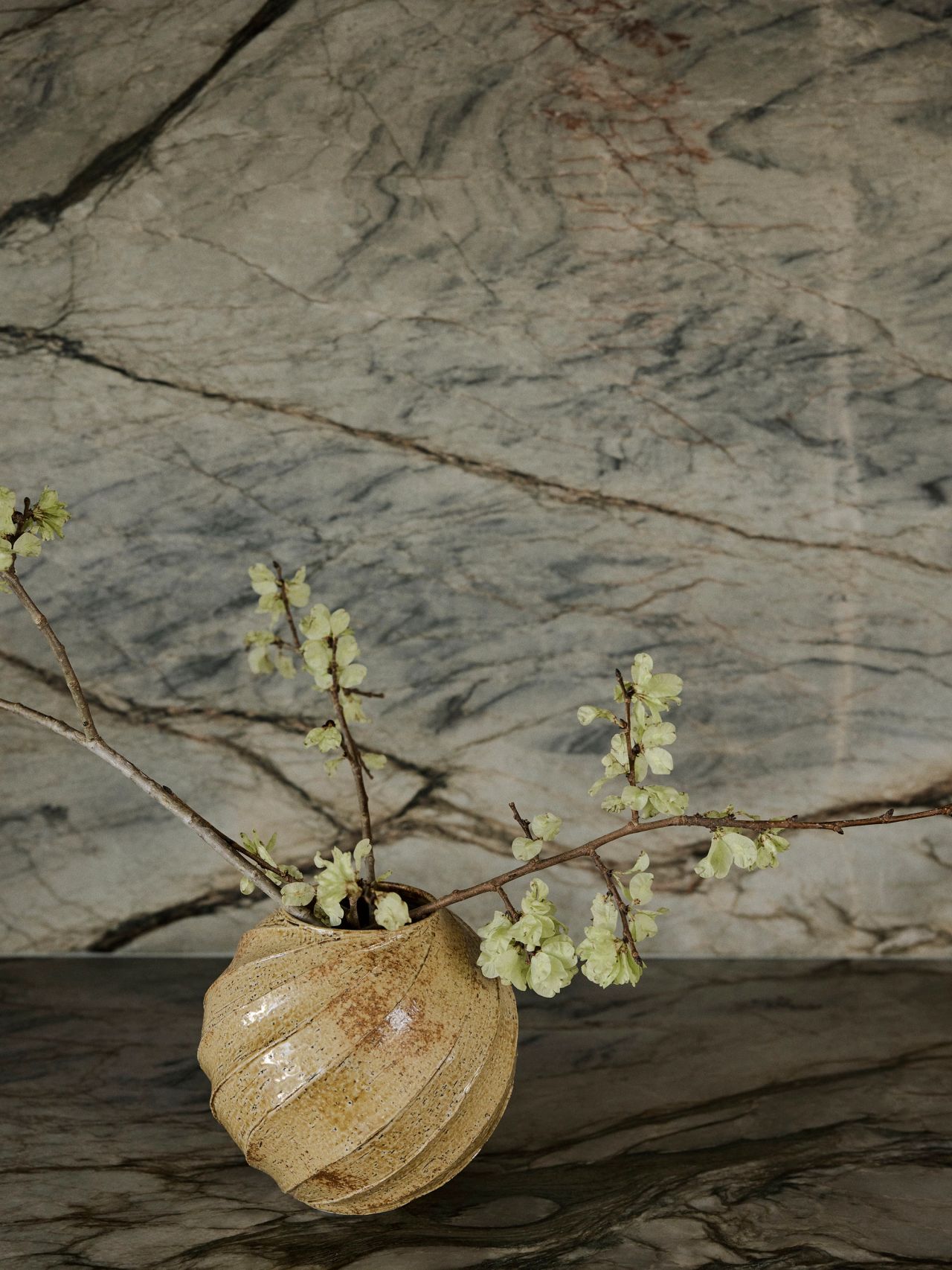 Sunset Ribbed ceramic vase with pale yellow-green flowering branches, set against a dramatic grey-veined marble backdrop