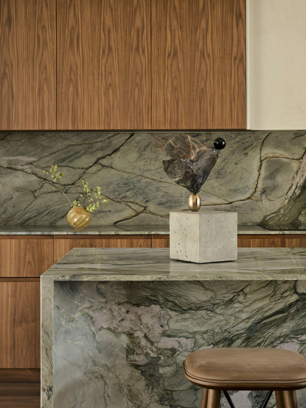 Sunset Sculptural bronze and stone object displayed on a concrete plinth atop a book-matched marble kitchen island, with a small textured vase and marble backsplash against walnut cabinetry.
