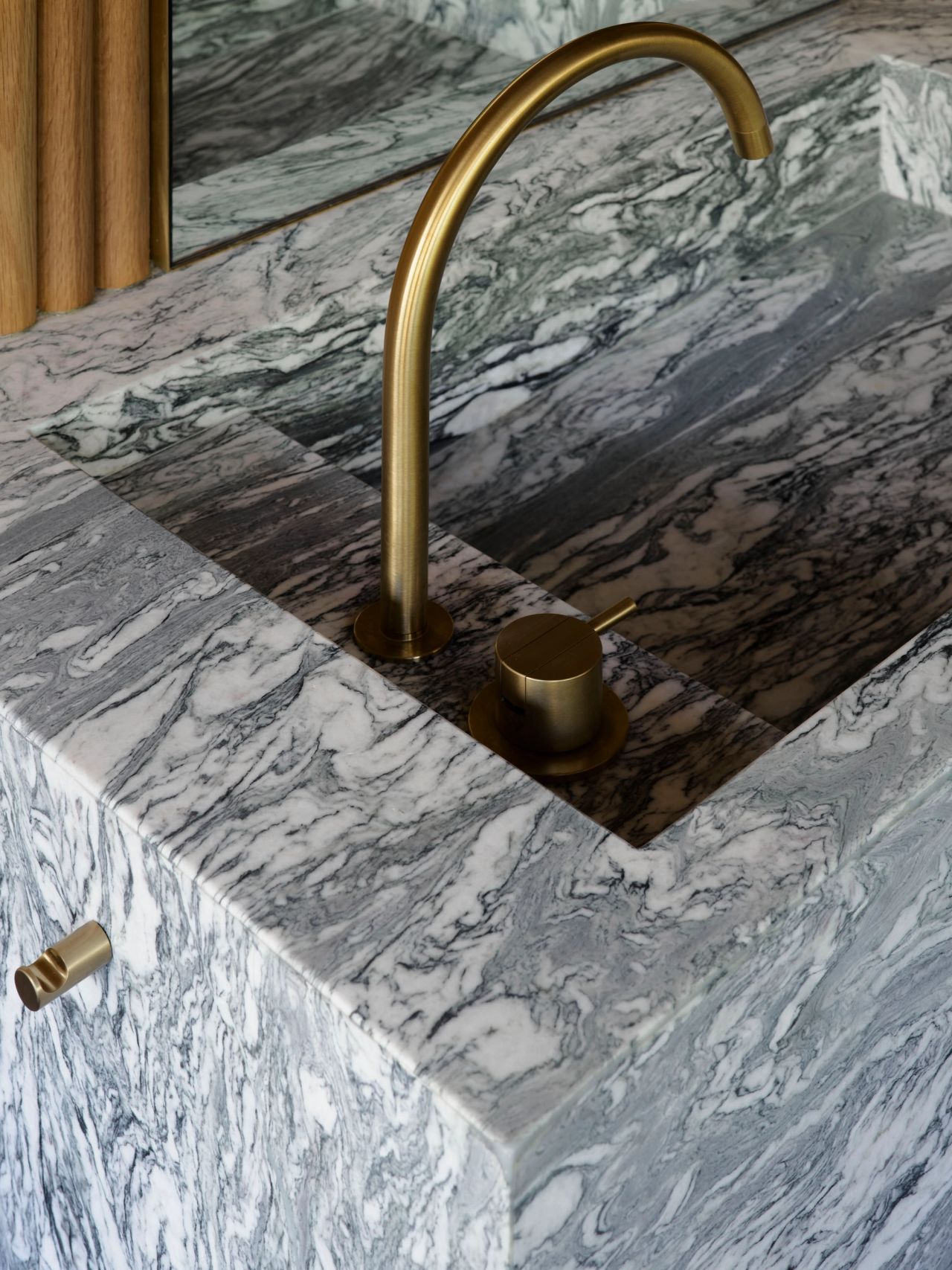 Ultramarine A close-up detail of a sculptural grey-and-white veined marble sink basin with a curved brushed-brass gooseneck tap and matching round single-lever mixer. The marble's dramatic swirling grain is echoed in a mirror reflection above, with a timber-panelled wall visible at the edge of the frame.