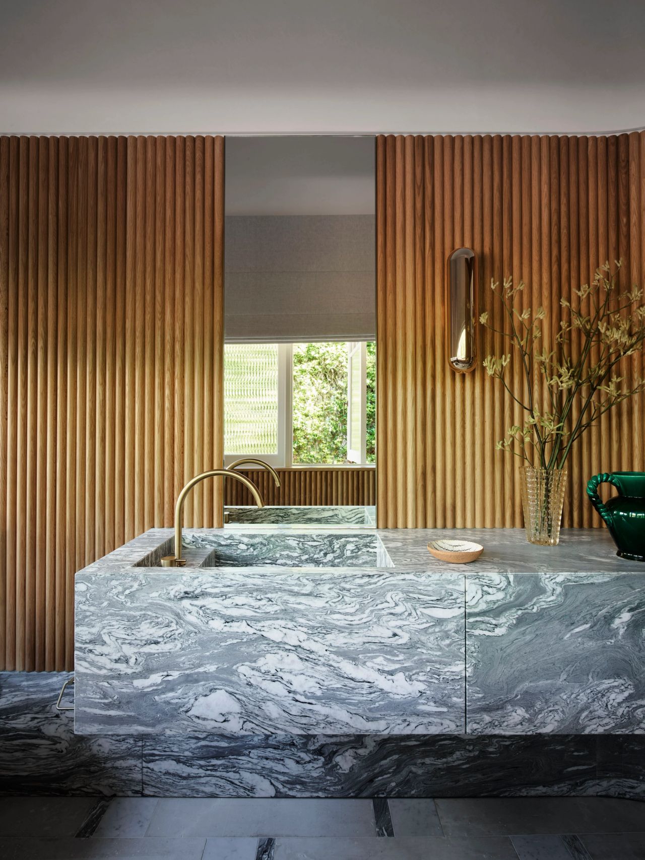Ultramarine A striking bathroom vanity carved from bold grey-and-white swirling marble with an integrated sink and brass gooseneck tap, set against a wall of vertical fluted timber slats. A cylindrical brass wall sconce is mounted to the right, above a glass vase of dried grasses and a green ceramic vessel on the counter. A narrow window with a woven roller blind sits centered between the timber panels, framing a view of greenery outside.