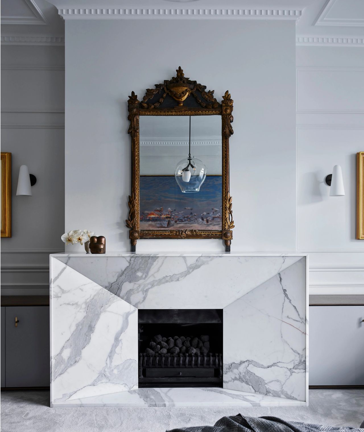 Ornate antique gilt mirror above a sculptural white marble fireplace, framed by pale grey panelled walls and contemporary wall sconces.