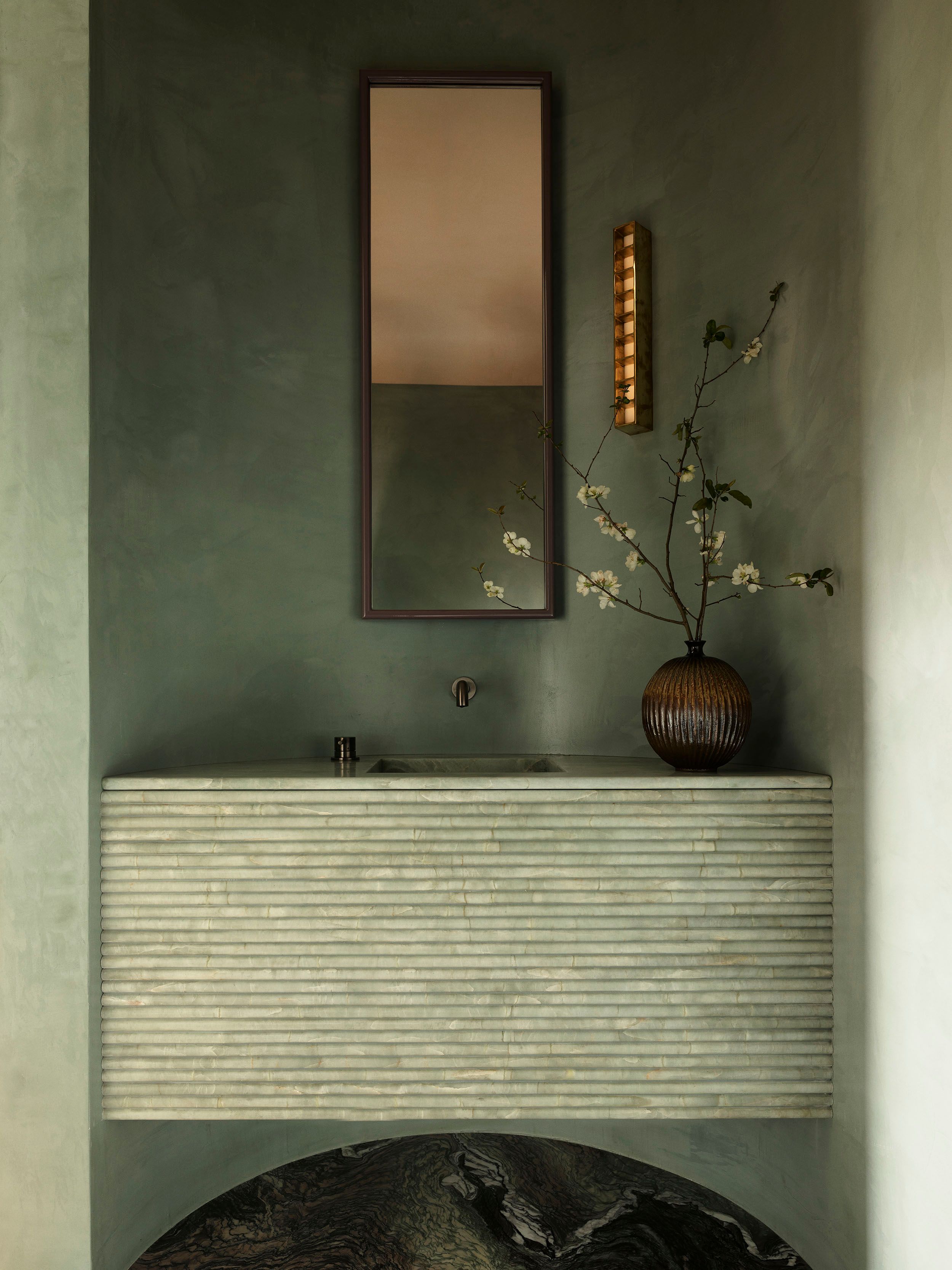 Sunset Ribbed stone vanity with integrated basin set against sage-green plaster walls, beneath a slim rectangular mirror and a ridged wall sconce, with a dark ribbed vase holding white blossom branches