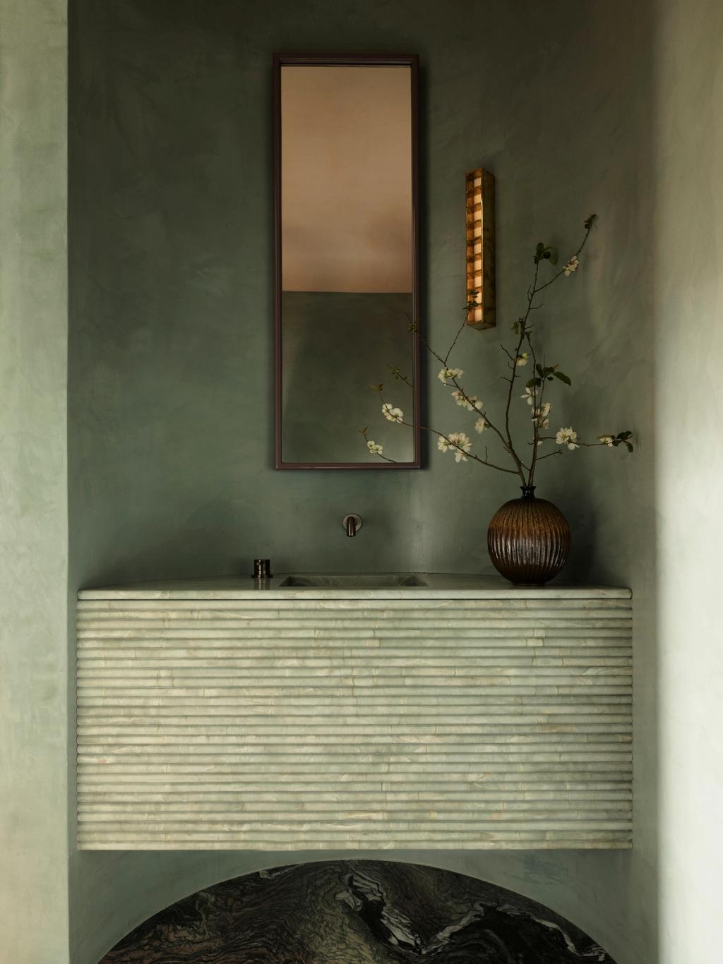 Sunset Ribbed stone vanity with integrated basin set against sage-green plaster walls, beneath a slim rectangular mirror and a ridged wall sconce, with a dark ribbed vase holding white blossom branches