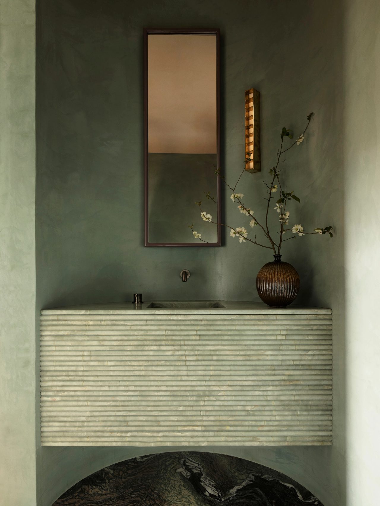 Sunset Ribbed stone vanity with integrated basin set against sage-green plaster walls, beneath a slim rectangular mirror and a ridged wall sconce, with a dark ribbed vase holding white blossom branches