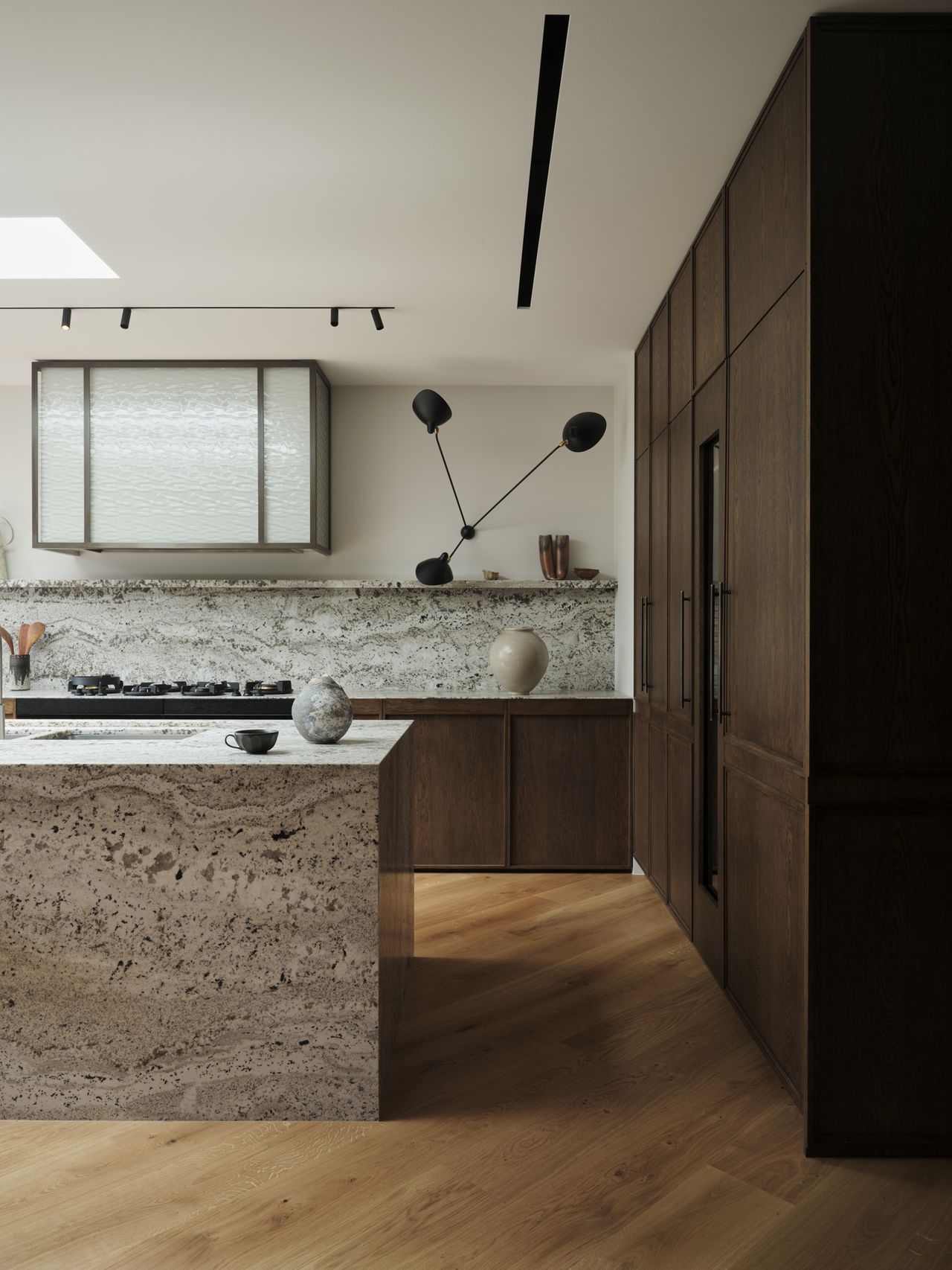 Monsieur G A modern kitchen with dark wood cabinetry and a textured stone countertop and waterfall island in warm beige travertine-like tones. A sculptural black articulated wall lamp with two angled arms hangs above the counter, near a fluted glass-front upper cabinet. A round ceramic sphere and a small bowl sit on the counter, with a gas cooktop set into the stone backsplash. Light oak flooring and a linear skylight complete the space