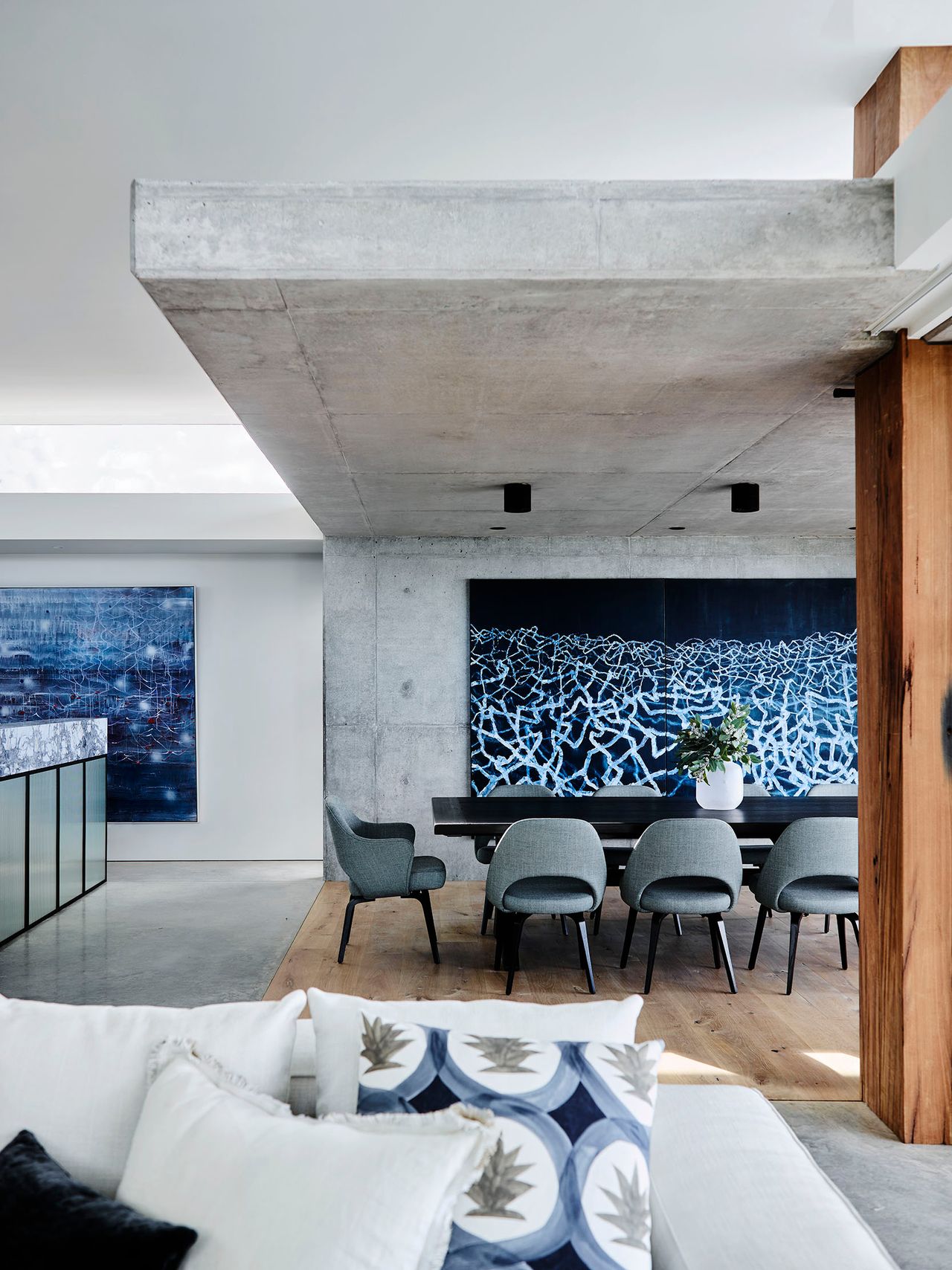Killcare Dining area beneath an exposed concrete ceiling beam, with a dark timber table and grey upholstered chairs against a large abstract navy artwork, and a white sectional sofa with patterned cushions in the foreground
