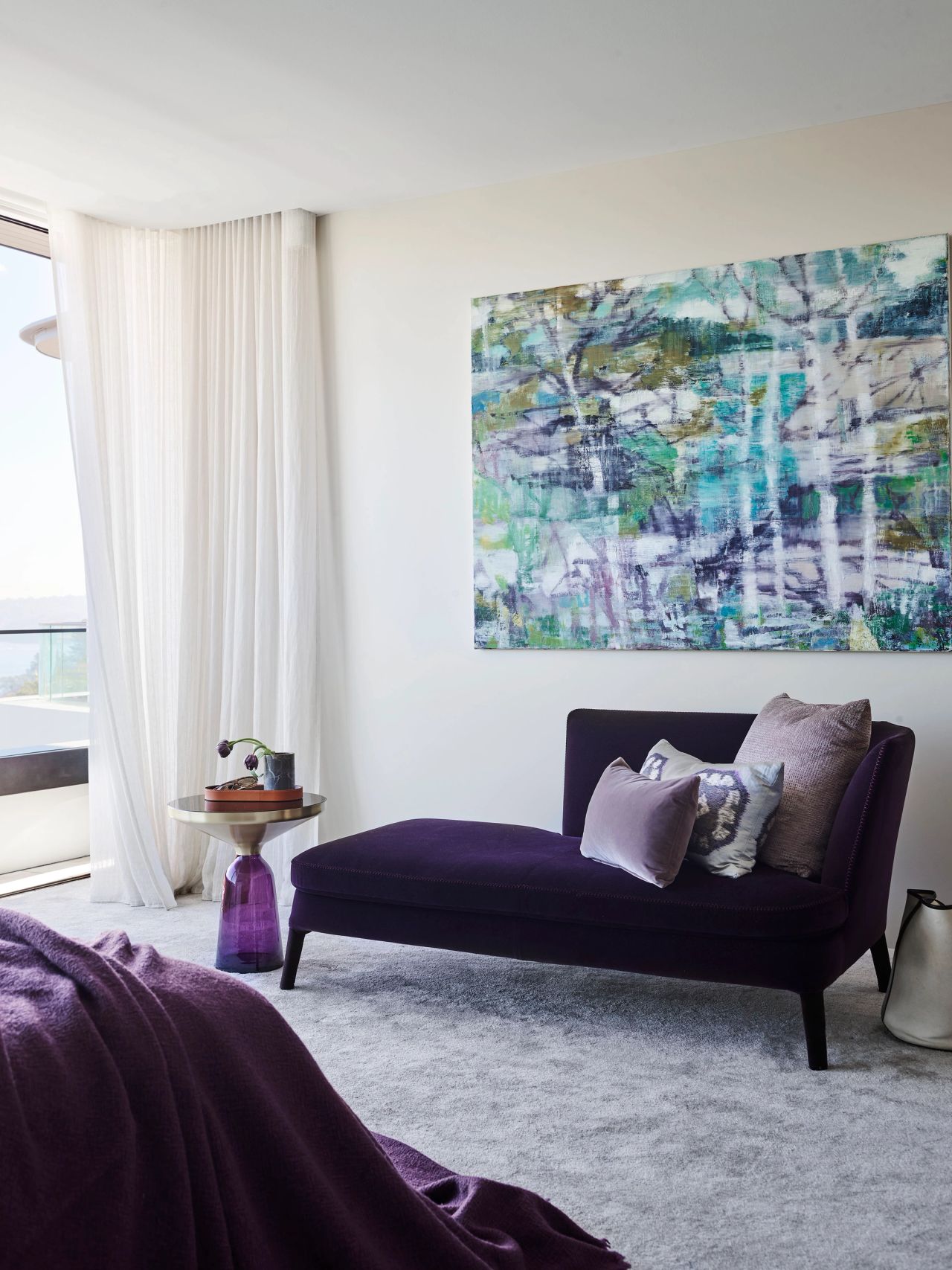 Contemporary bedroom seating area with deep purple chaise lounge, abstract artwork, sheer curtains and a sculptural side table.