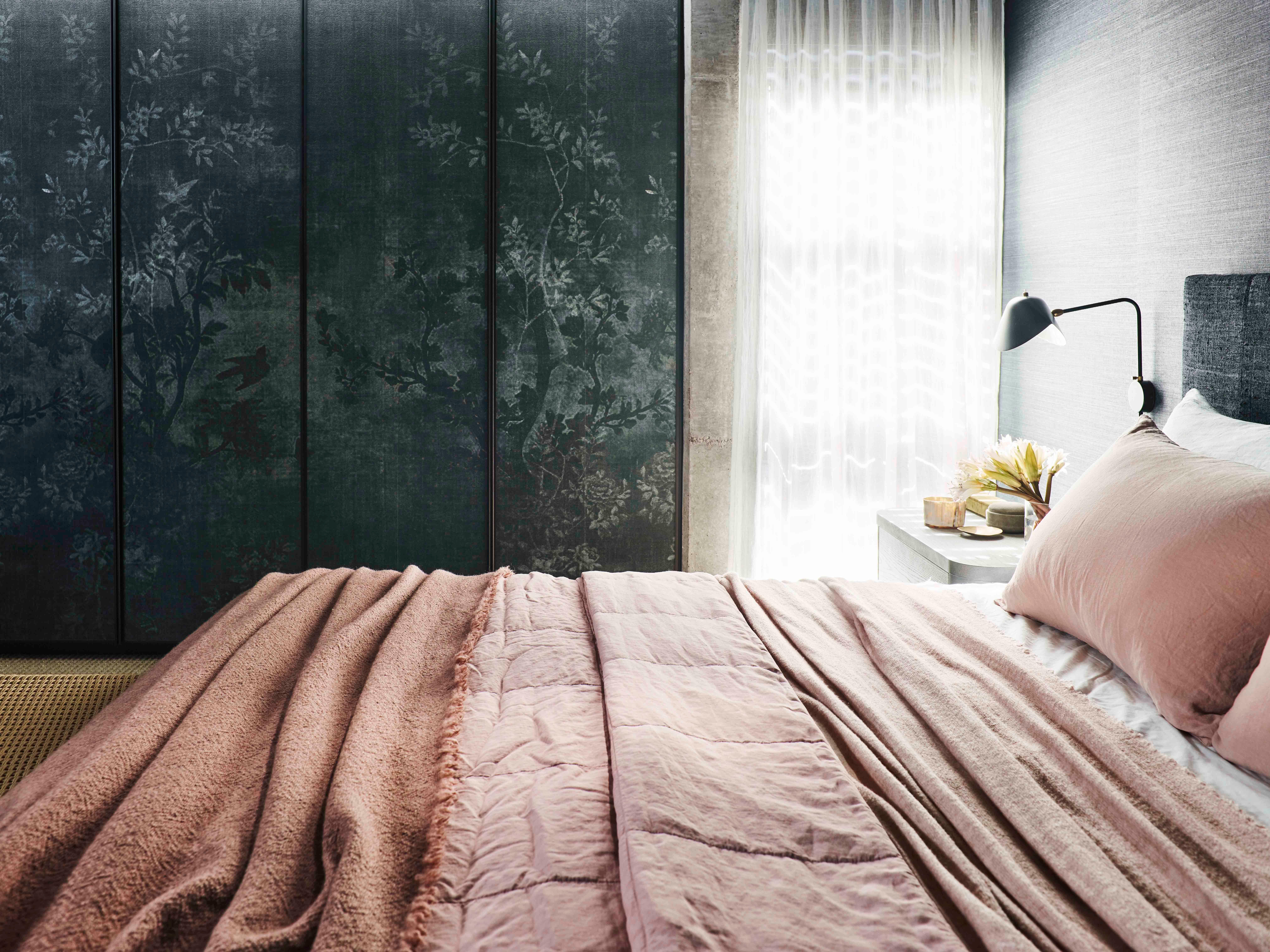 Killcare Bedroom detail with blush pink quilted linen bedding, a black articulated wall lamp above a stone bedside table styled with flowers and candles, and a dark floral chinoiserie wardrobe panel beside sheer curtains