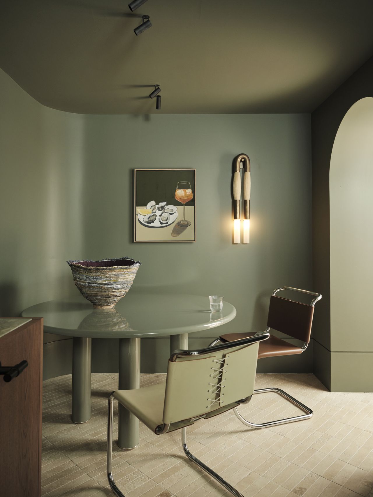 Monsieur G Sage-green cellar space with a curved round table on cylindrical legs, chrome-framed leather dining chairs, a large ceramic vessel as tabletop styling, a still-life oyster painting, and a vertical double-flame wall sconce
