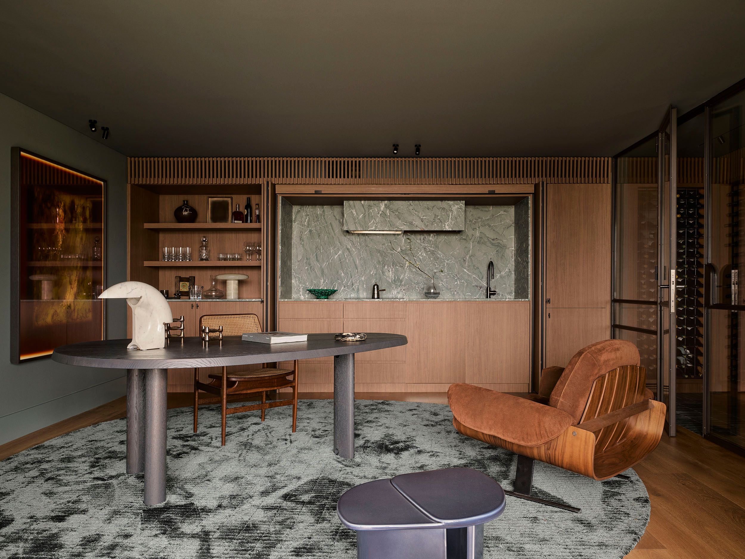 Sunset Study and bar area with a dark oval table on sculptural legs, a curved leather and wood lounge chair, a built-in wet bar with marble backsplash, and open wood shelving displaying glassware, set on a mottled charcoal rug