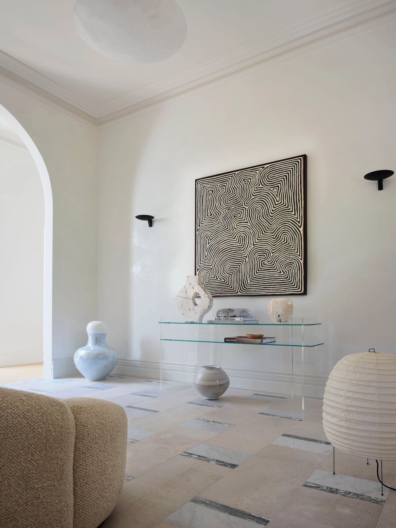 Ultramarine A pale, minimalist living space with a large abstract black-and-white maze-patterned artwork centered on the wall above a slim glass console table. Two black disc-shaped wall sconces flank the artwork. The console holds an abstract sculptural object, stacked books, and a small ceramic vessel. A pale blue bulbous ceramic vase and a grey rounded vase sit on the patterned stone floor, alongside a round white paper globe lamp on thin black legs. A beige boucle sofa is visible in the foreground, and an arched doorway opens to the left.