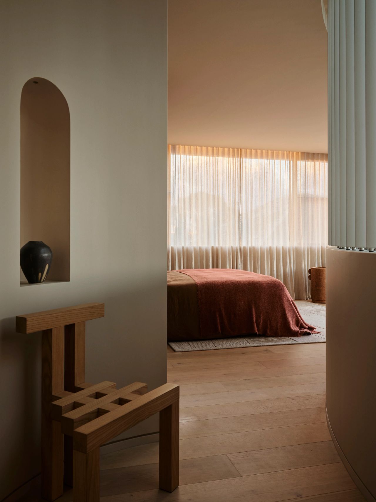 Patchwork Close-up of a wave-edged wooden bedside table with a small speckled ceramic vase, beneath a brass reading light mounted on a herringbone-textured headboard, next to a rust-coloured bed.
