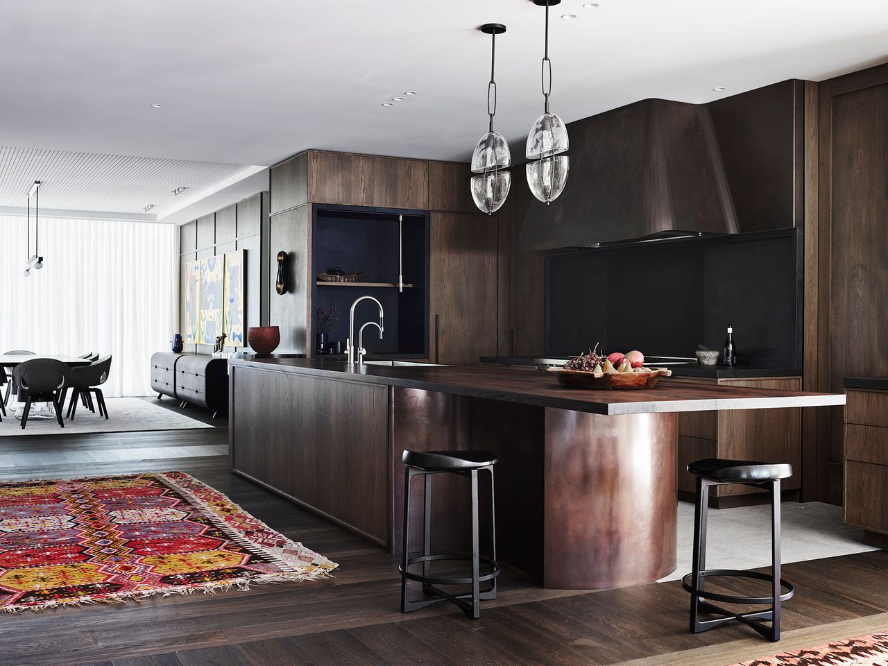 Contemporary kitchen with dark timber cabinetry, a sculptural bronze island, glass pendants and richly patterned rugs, balancing refined materials with warmth and character.