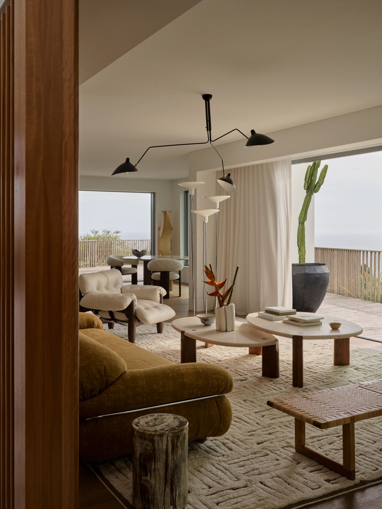 Dancing on the Ceiling Light-filled coastal living room with a sculptural multi-arm black wall lamp, round nested coffee tables, an olive velvet sofa, and a tall potted cactus, opening onto a terrace with ocean views