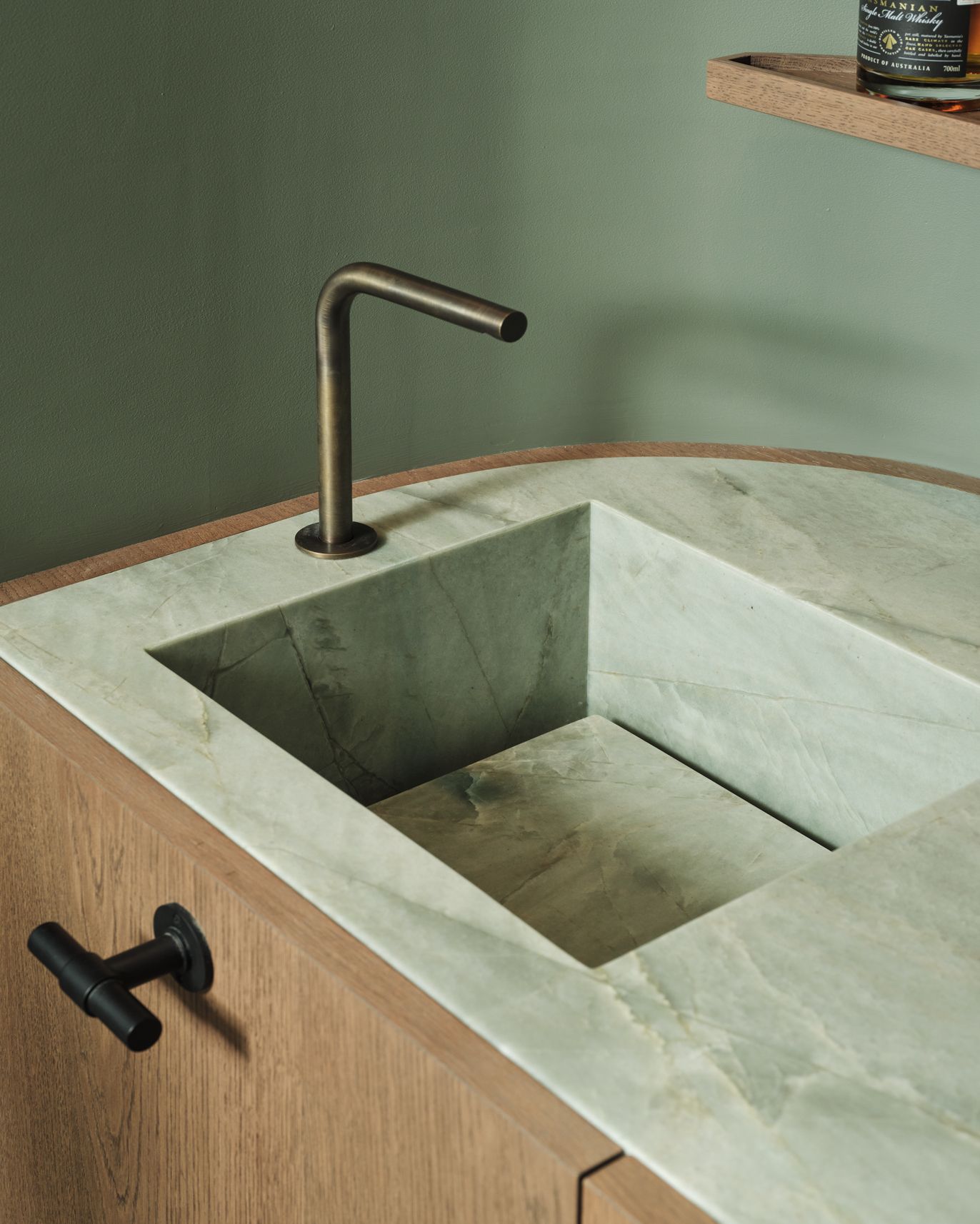 Monsieur G Detail of a bar sink carved from pale green-toned stone with an angular brass tap, set into a curved timber cabinet with a black cylindrical drawer pull