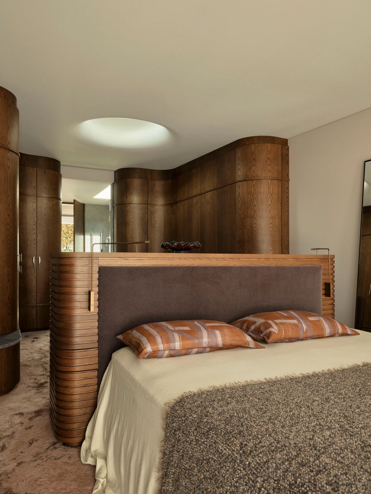 Sunset Bedroom with a curved, ribbed wood bedhead structure and integrated side units, an upholstered headboard, patterned throw pillows, and a circular skylight above wardrobe cabinetry beyond