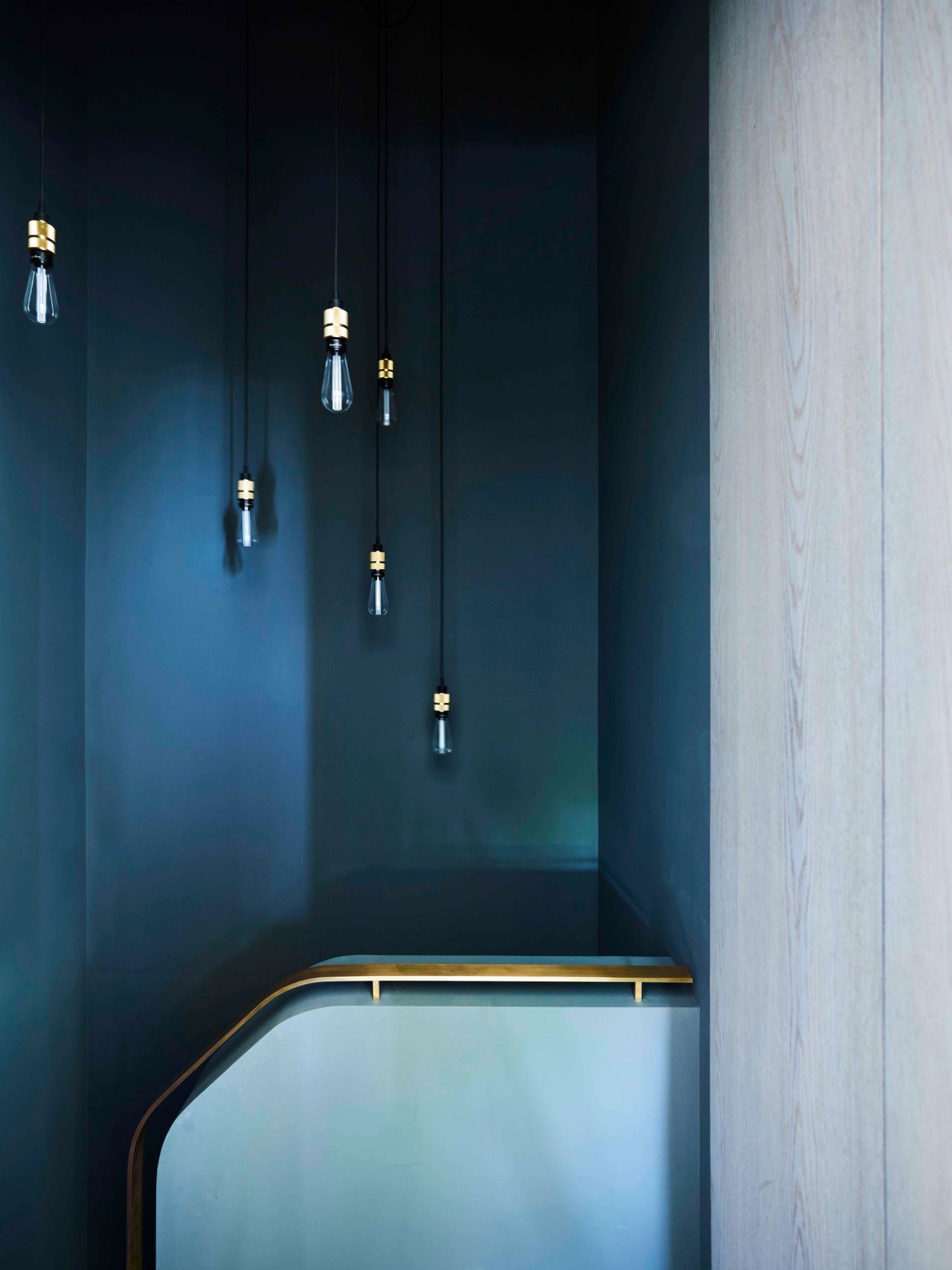 Killcare Cluster of exposed-bulb pendant lights hanging at varying heights above a curved brass-edged stair balustrade, against a dark navy stairwell wall