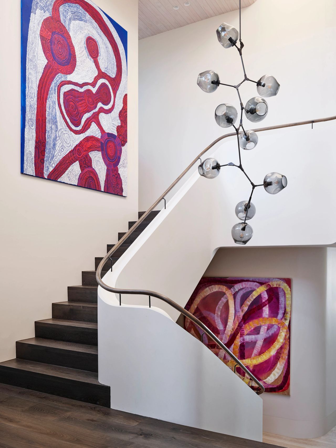 Hill House staircase curved rendered balustrade with metal handle. one large indigenous work of blue, pink and white and second colourful abstract artwork. Big glass Smokey grey pendant  