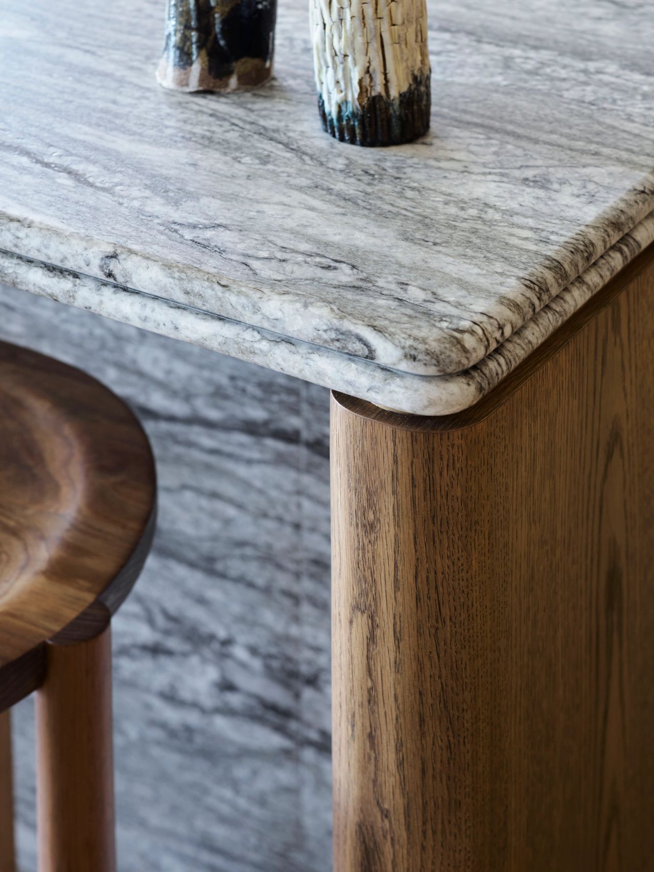 Ultramarine Close-up of a rounded grey marble countertop edge meeting a curved wooden corner post, with small ceramic vases on top and a wooden stool beside it